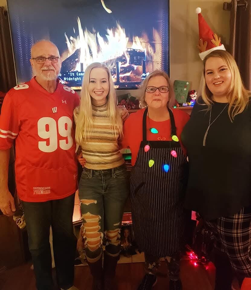 Four people pose together indoors in front of a lit fireplace with holiday decorations and a Christmas hat on the rightmost person.