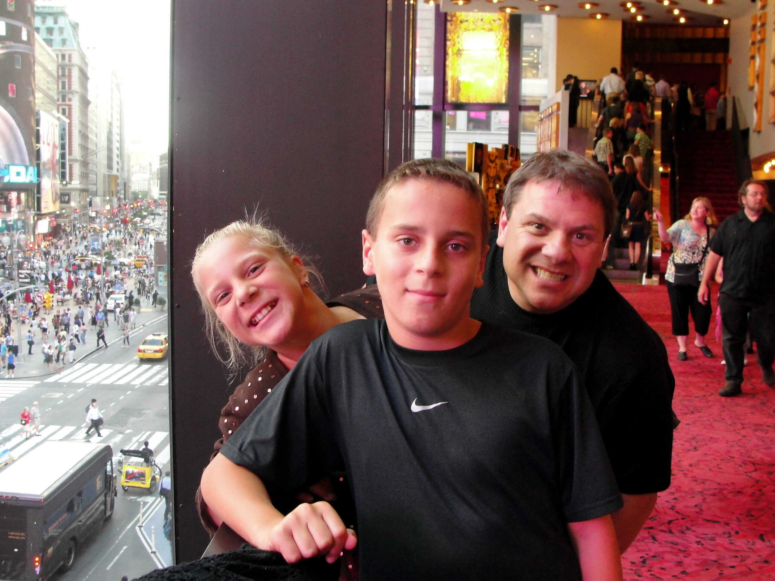 Three friends posing for a selfie in a busy indoor lobby, with a city street visible through large windows on the left.