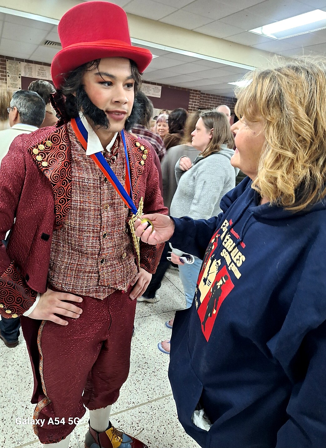 Young person wearing a red Victorian costume with a top hat and medals greets a woman at a crowded event.