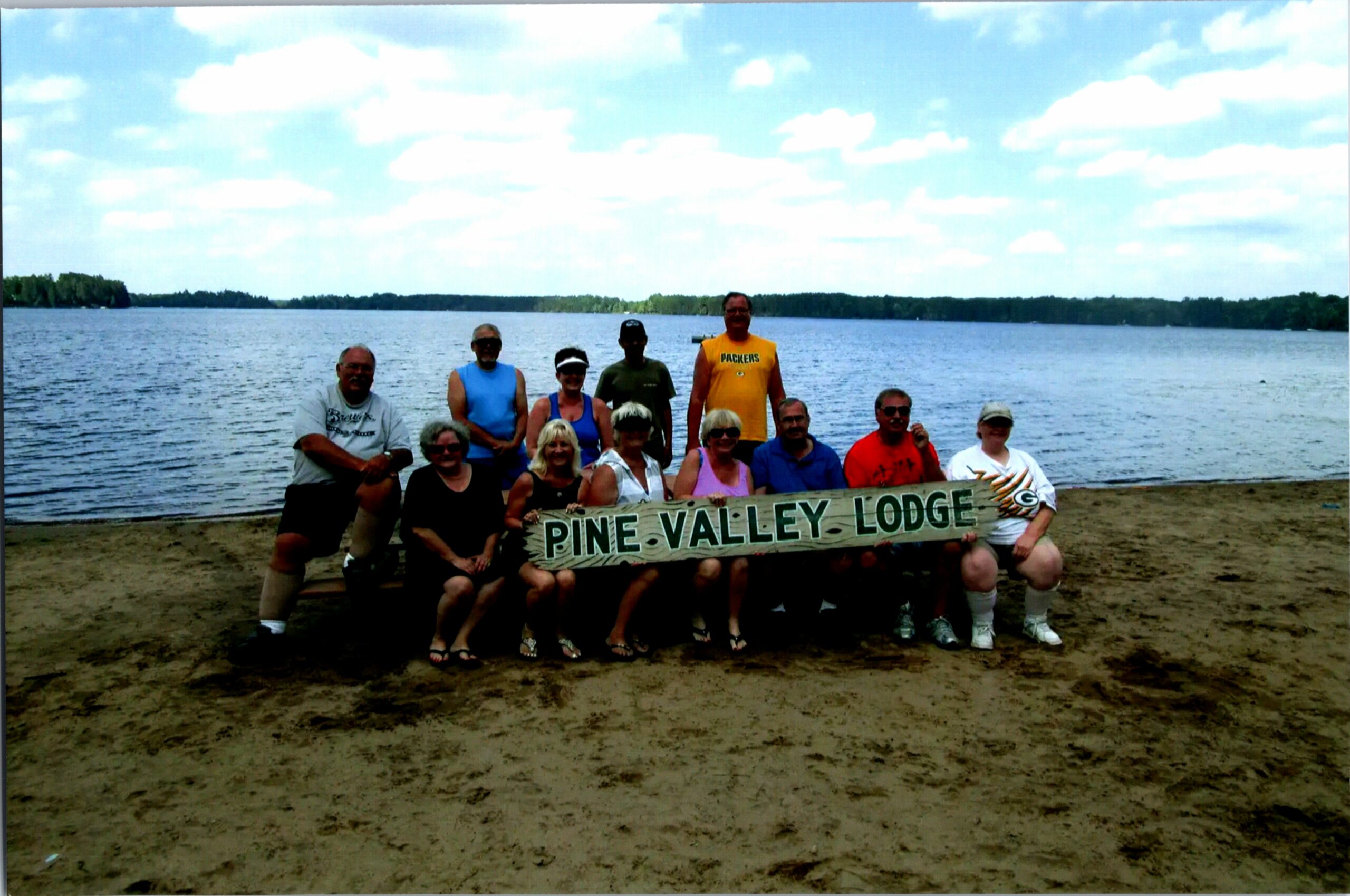 Group of people on a sandy lakeshore holding a banner that reads Pine Valley Lodge, with a calm lake and trees in the background.