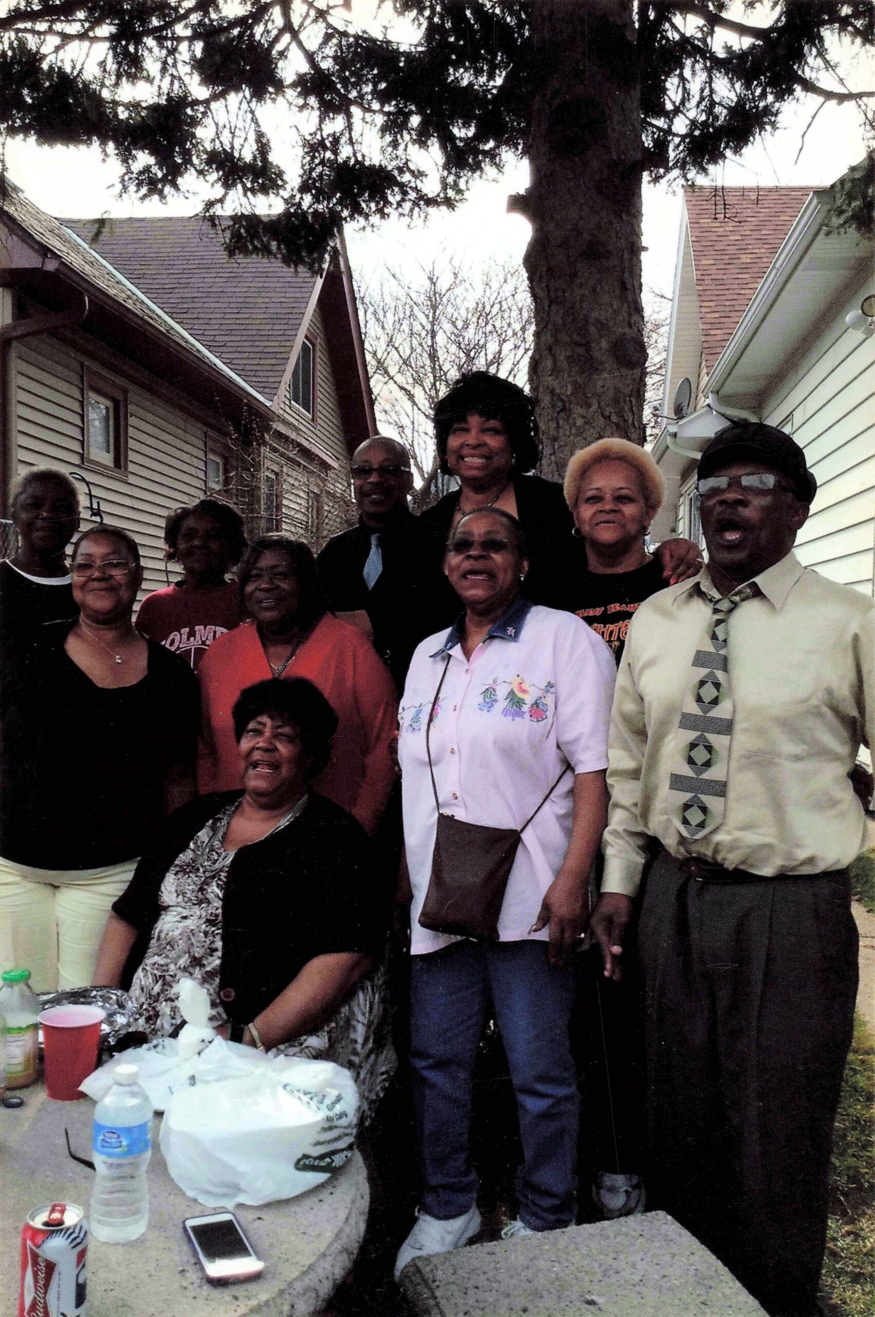 A group of adults posing and smiling outdoors in a residential backyard with a large tree behind them.