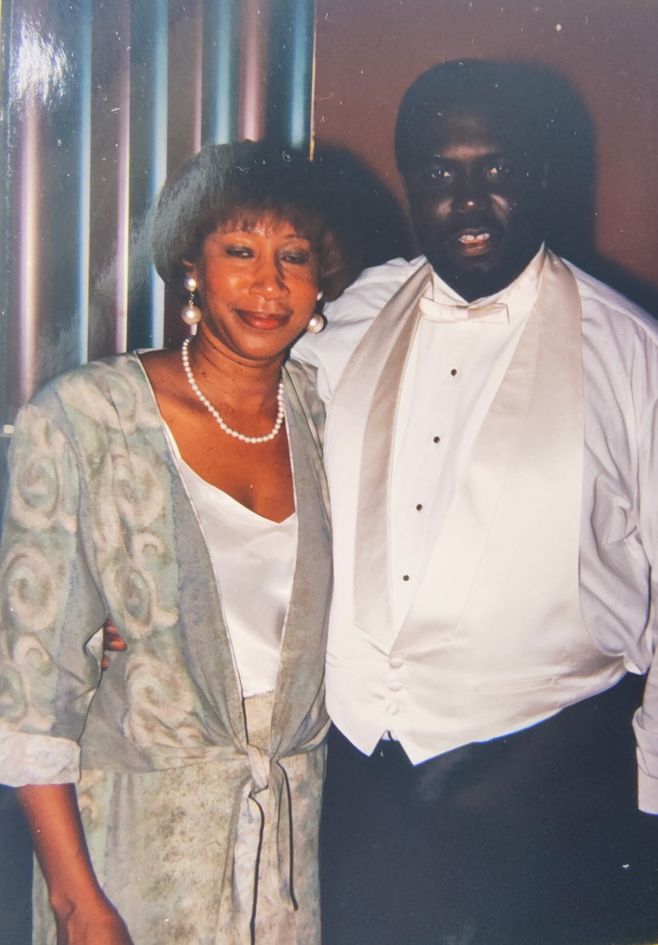 Smiling couple in formal wear posing together; woman wears a patterned robe with pearls, man wears a white tuxedo and vest with a bow tie on a striped backdrop.