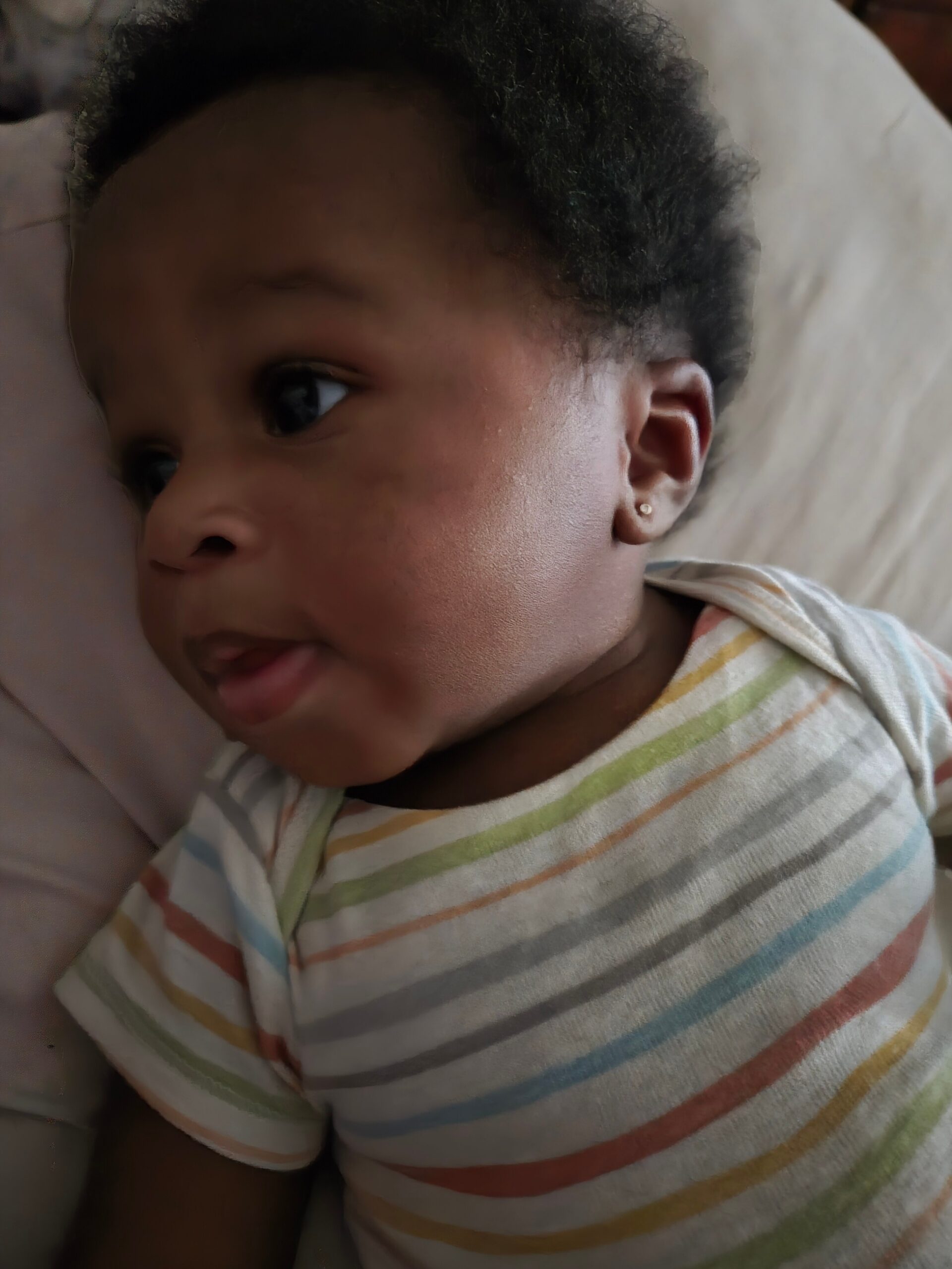 Close-up of a baby with dark curly hair wearing a pastel striped onesie, lying on a soft surface and looking to the side.