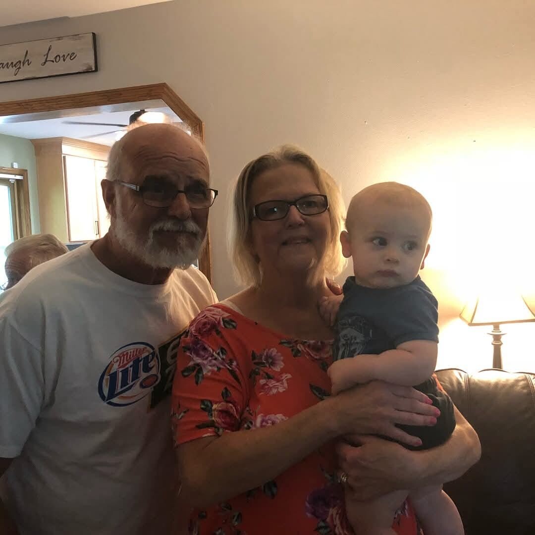 Elderly man with white beard and glasses stands next to a smiling woman in a red floral blouse, holding a baby in blue clothing indoors.