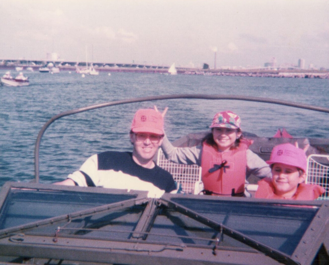 Man and two children in pink life jackets sit in a small motorboat, smiling at the camera with the water and harbor in the background.