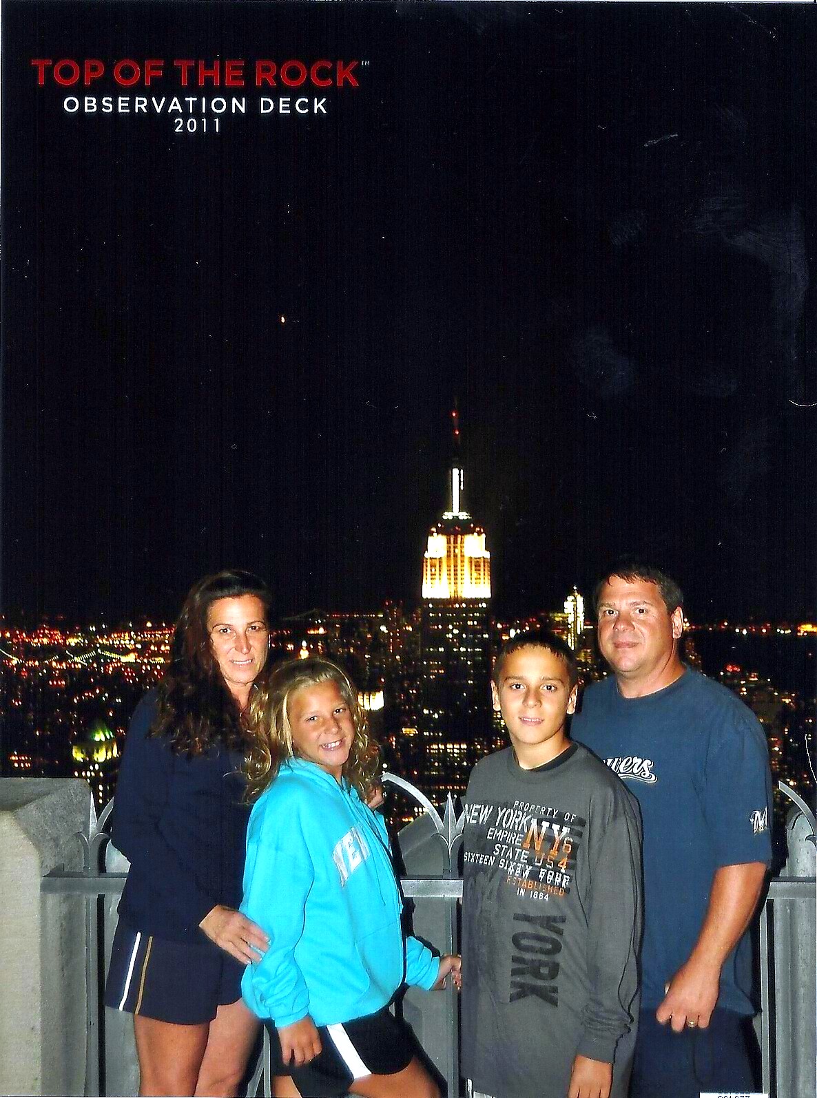 Group of four posing on a rooftop railing at night with the lit New York City skyline and the Empire State Building in the background.