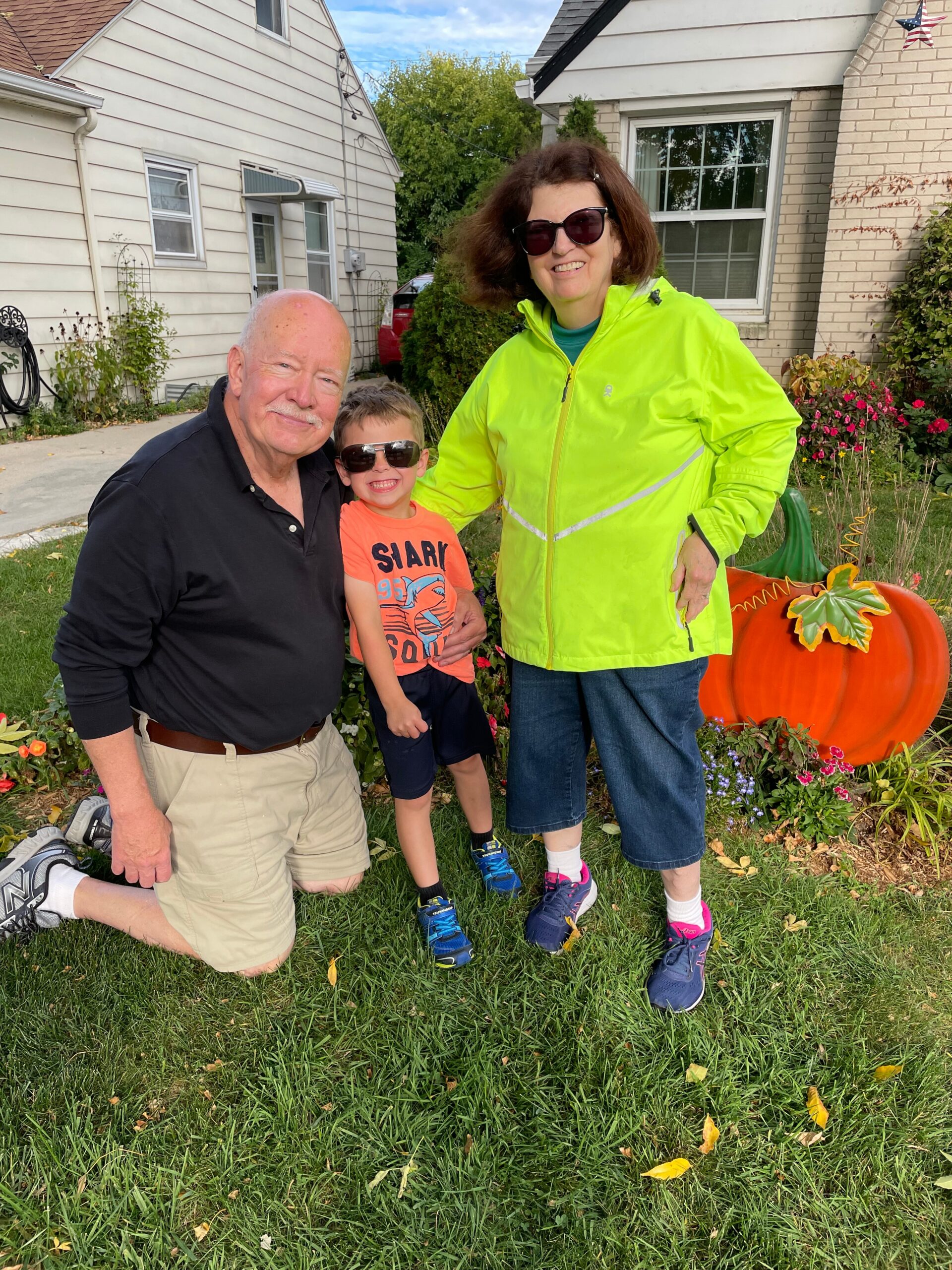 Smiling man, child, and woman in a bright jacket pose in a yard with a carved pumpkin display behind them.