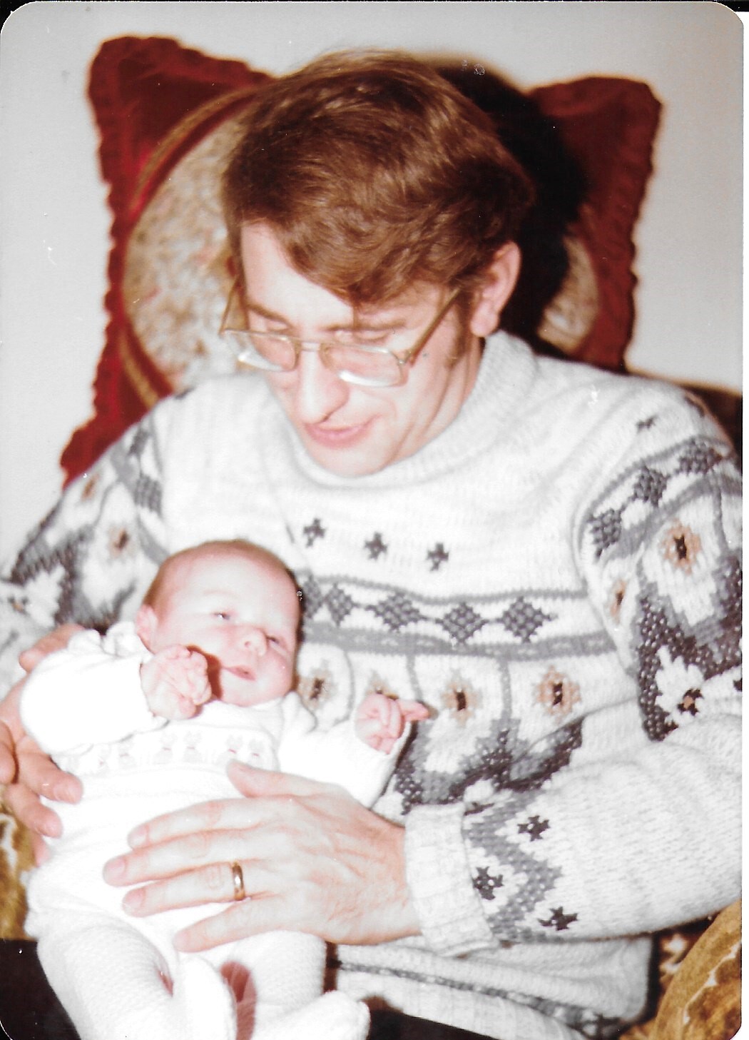 Older woman with glasses wearing a patterned sweater cradles a newborn baby in white clothing.