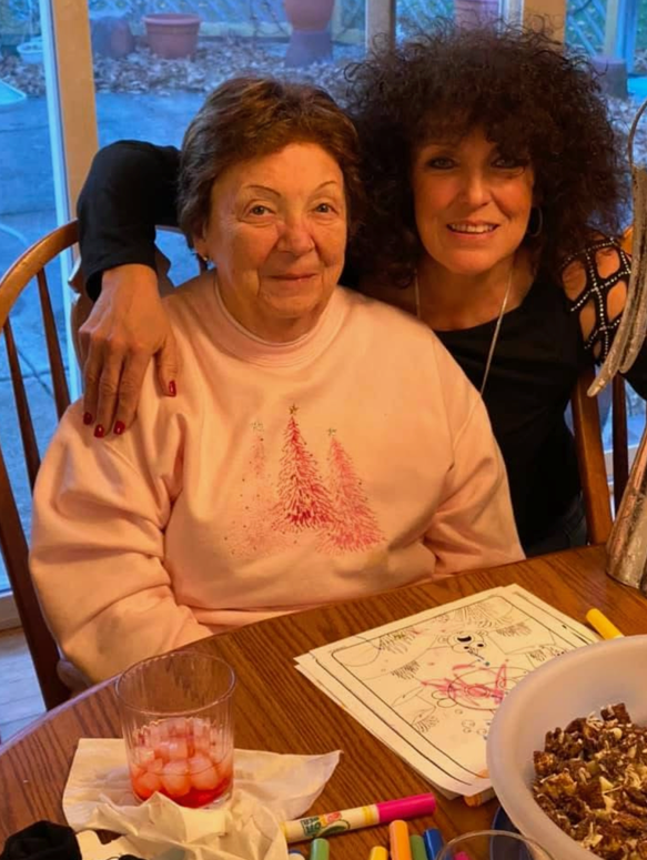 Two women sit close together at a wooden table indoors; an older woman in a pink sweater with a Christmas tree design sits beside a younger woman with dark curly hair, arm around her. On the table are coloring sheets, markers, a glass with ice, and a bowl of snacks; a windowed background appears behind them.