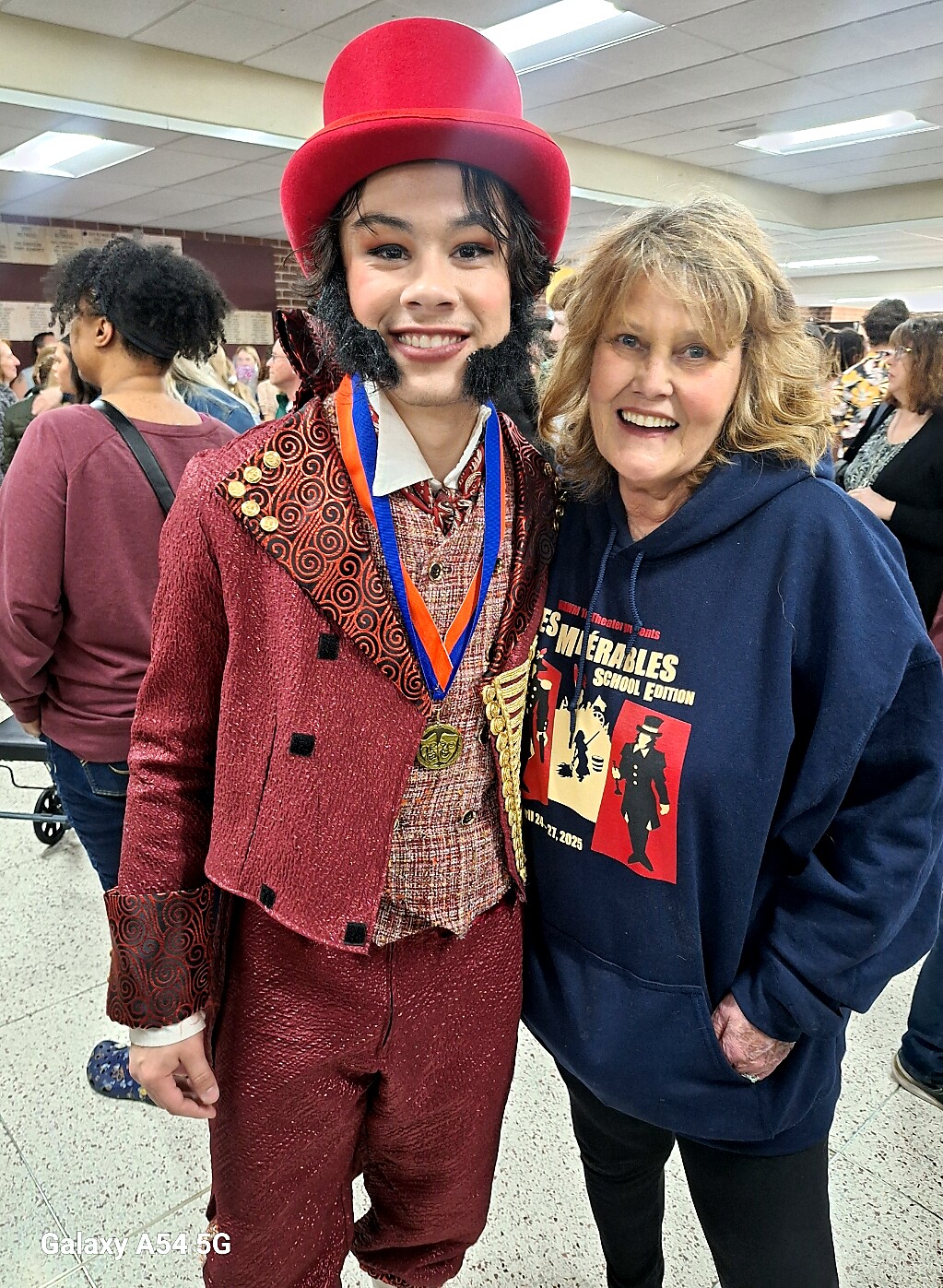 Young person in a red Willy Wonka-inspired costume with a tall red hat and medals posing with an smiling older woman in a navy hoodie at a crowded indoor event.
