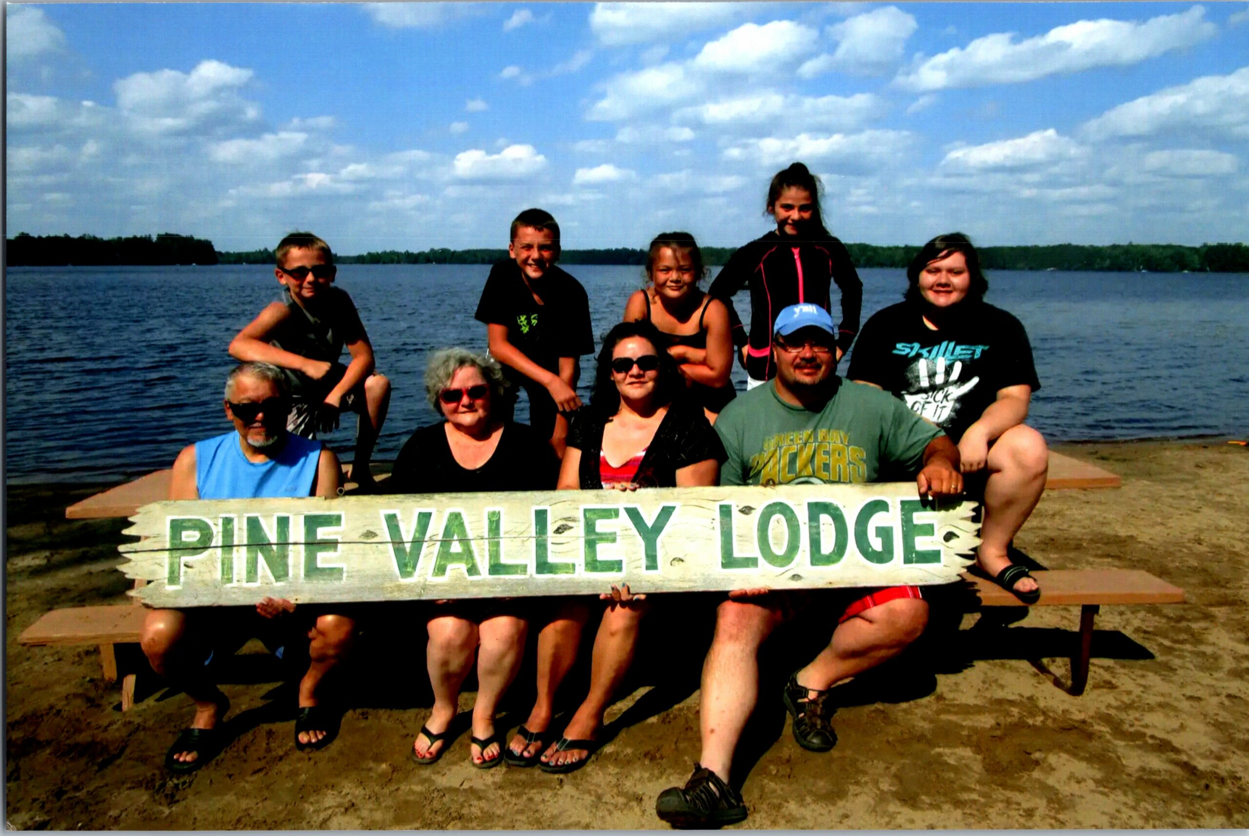 Group of people posing on a lakeside dock, holding a weathered sign that reads Pine Valley Lodge, with a blue lake and sky in the background.