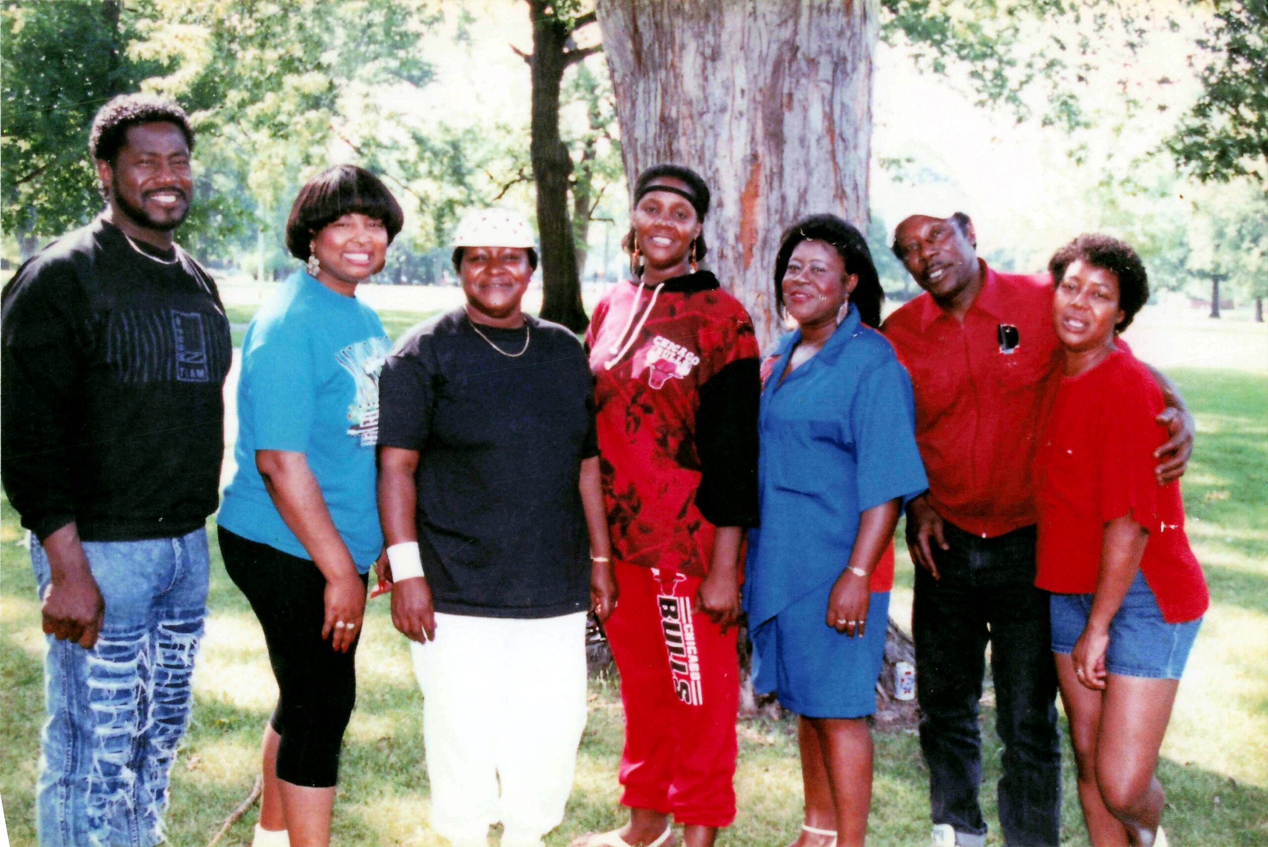 Group of seven adults posing in a park in front of a large tree, smiling at the camera.