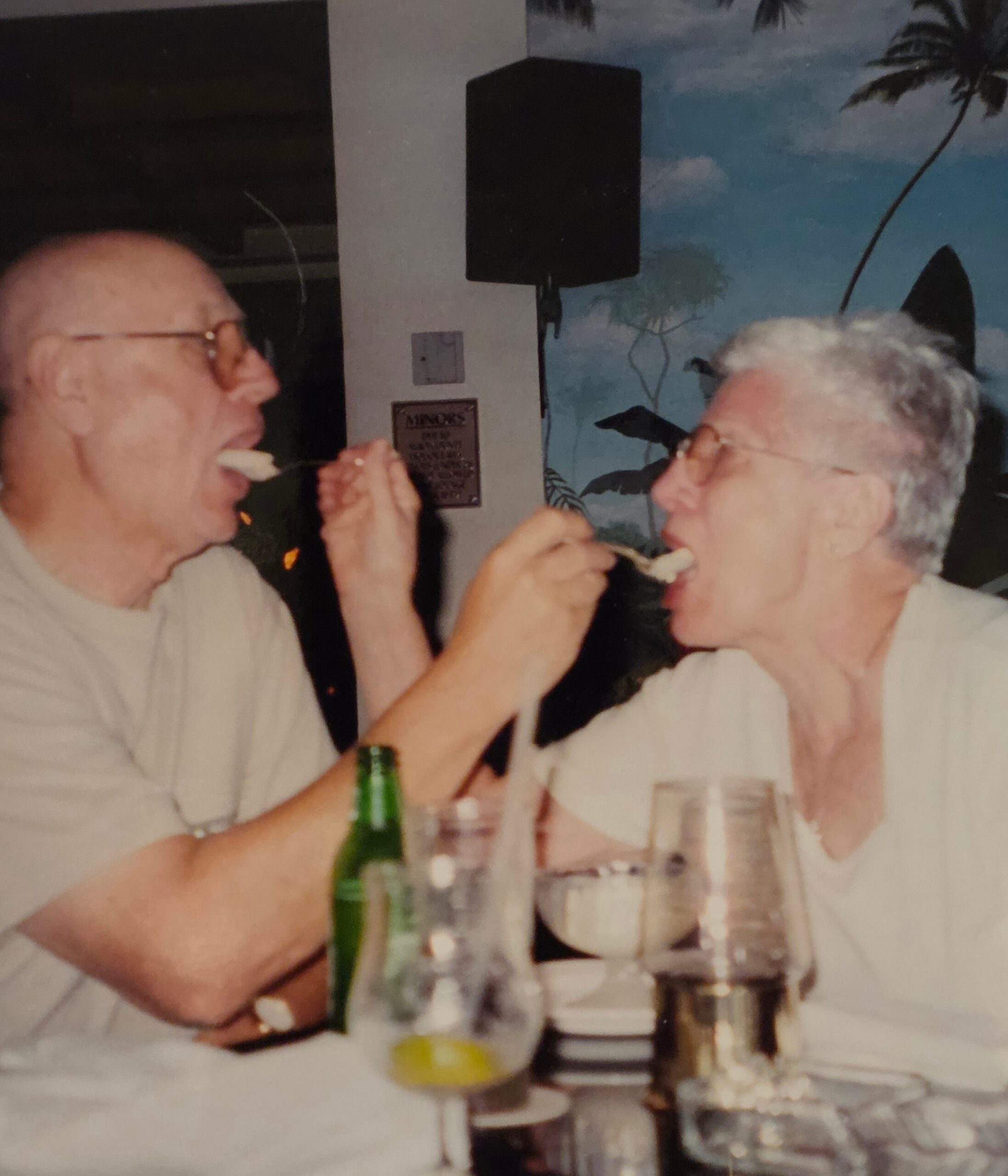 Two older men at a dining table share a bite, with a beer bottle and glasses in front of them.
