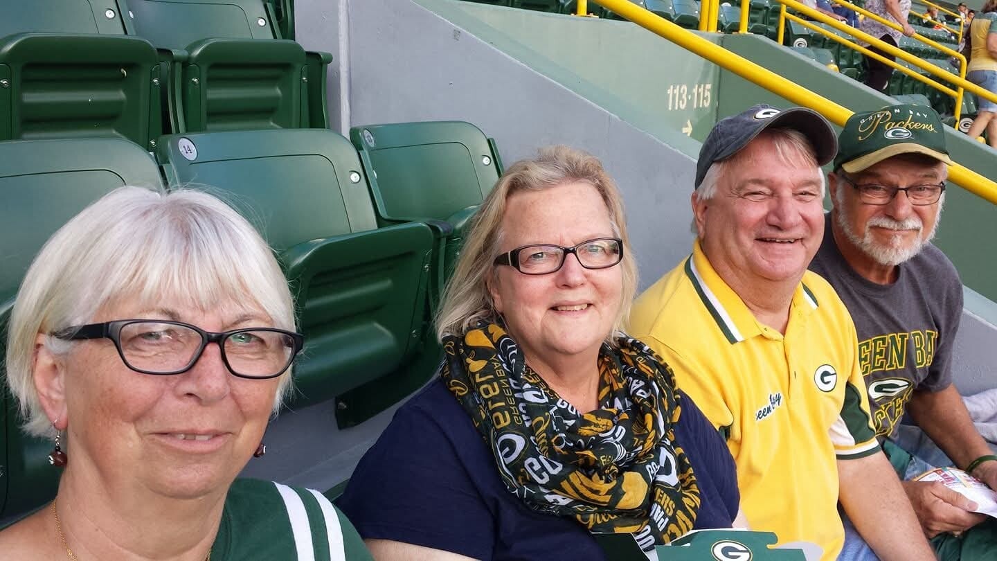 Four sports fans sit in green stadium seats, smiling for a selfie at a Packers game; two women in front, two men behind wearing team gear.