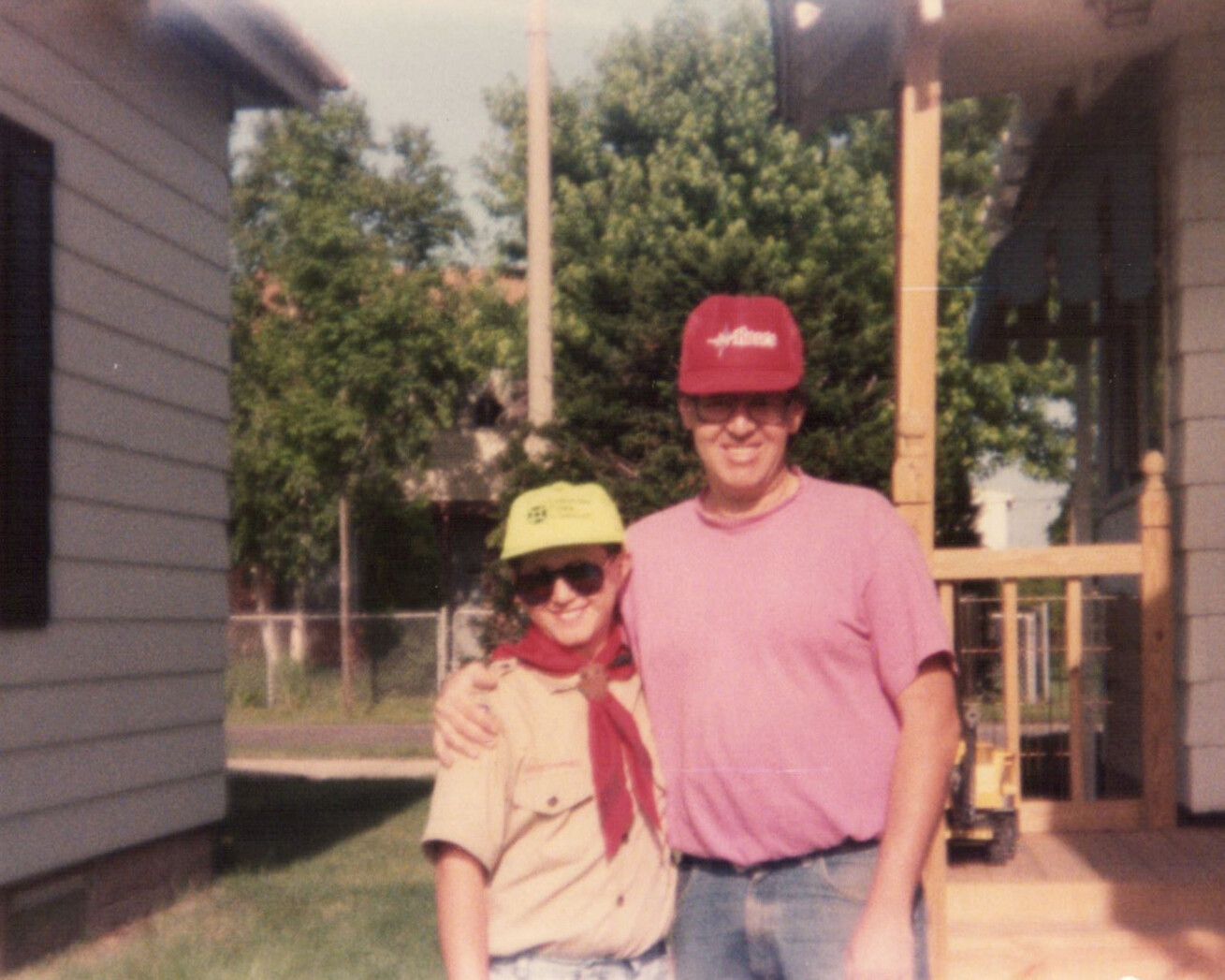 Two smiling people stand arm in arm in a backyard, wearing bright caps (yellow and red) near a wooden playset.