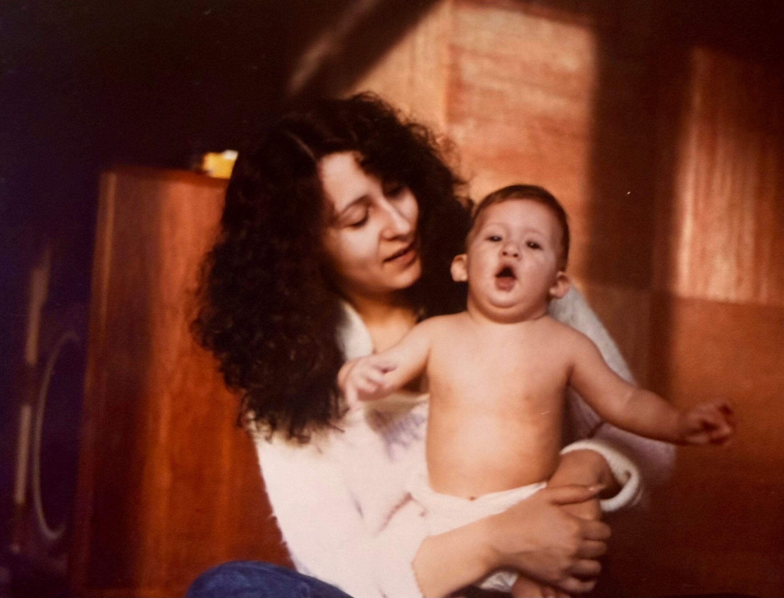Woman with curly hair holding a baby indoors against a wooden wall.