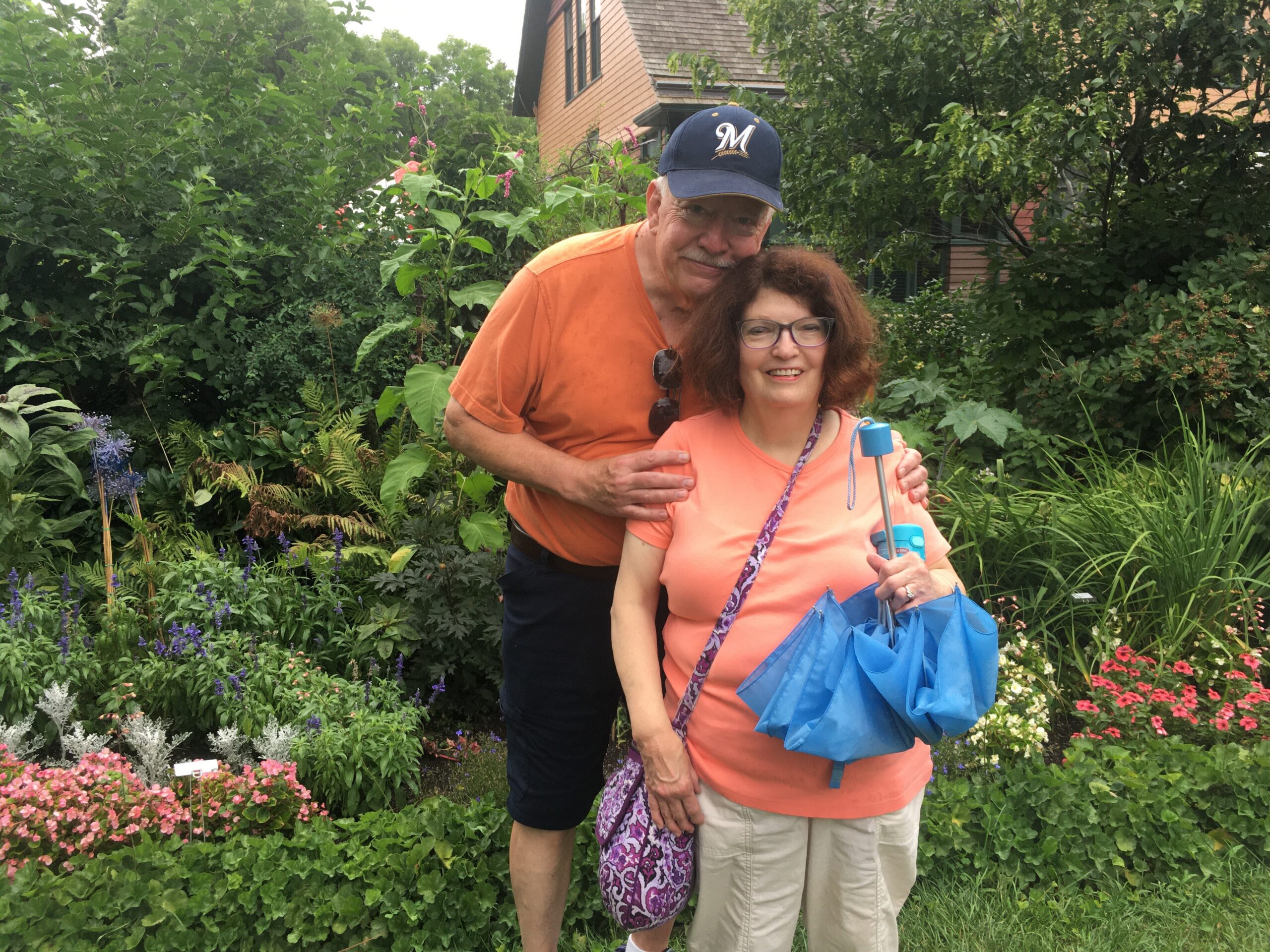 Smiling couple posing in a lush garden, the man in an orange shirt has his arm around the woman in an orange top, surrounded by flowers and greenery.
