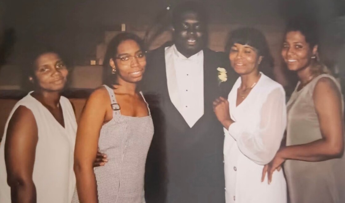 Five adults pose together in a formal setting; man in a tuxedo with boutonniere center, flanked by four women in dresses, smiling at the camera.
