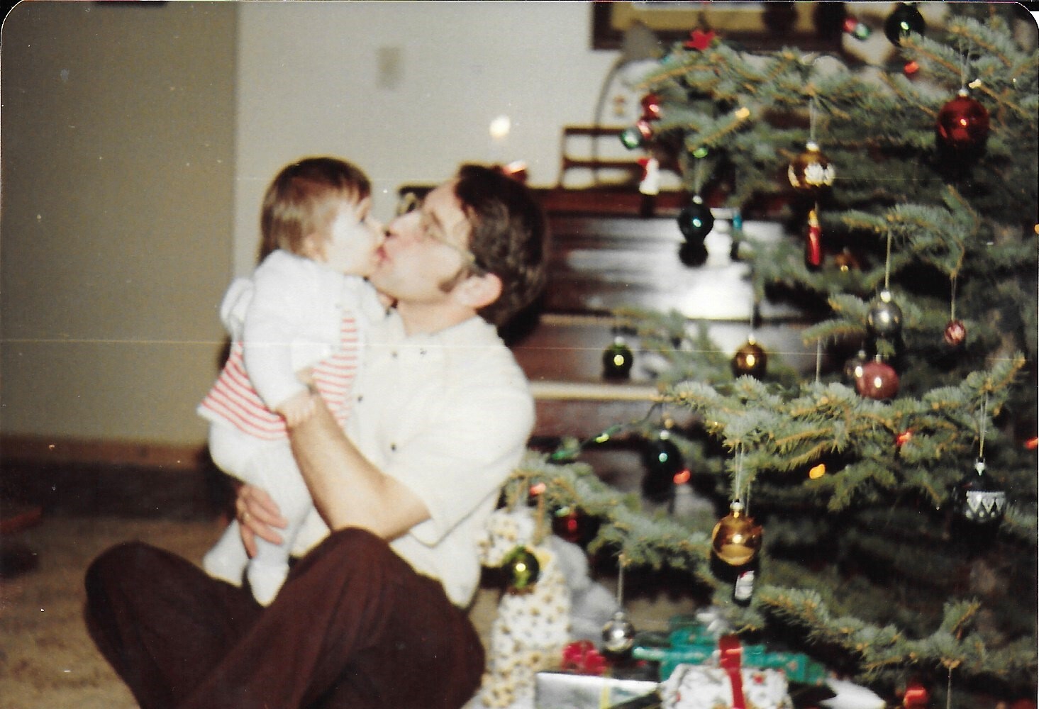 Father holding a baby and sharing a kiss beside a Christmas tree with ornaments and wrapped gifts nearby
