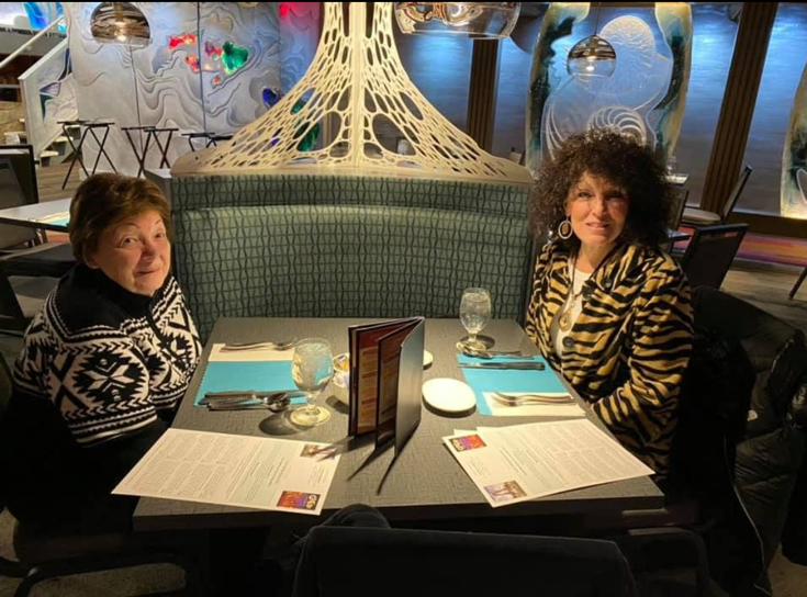 Two women sit across from each other at a restaurant table with menus, glassware, and turquoise placemats, a white sculptural centerpiece behind them.