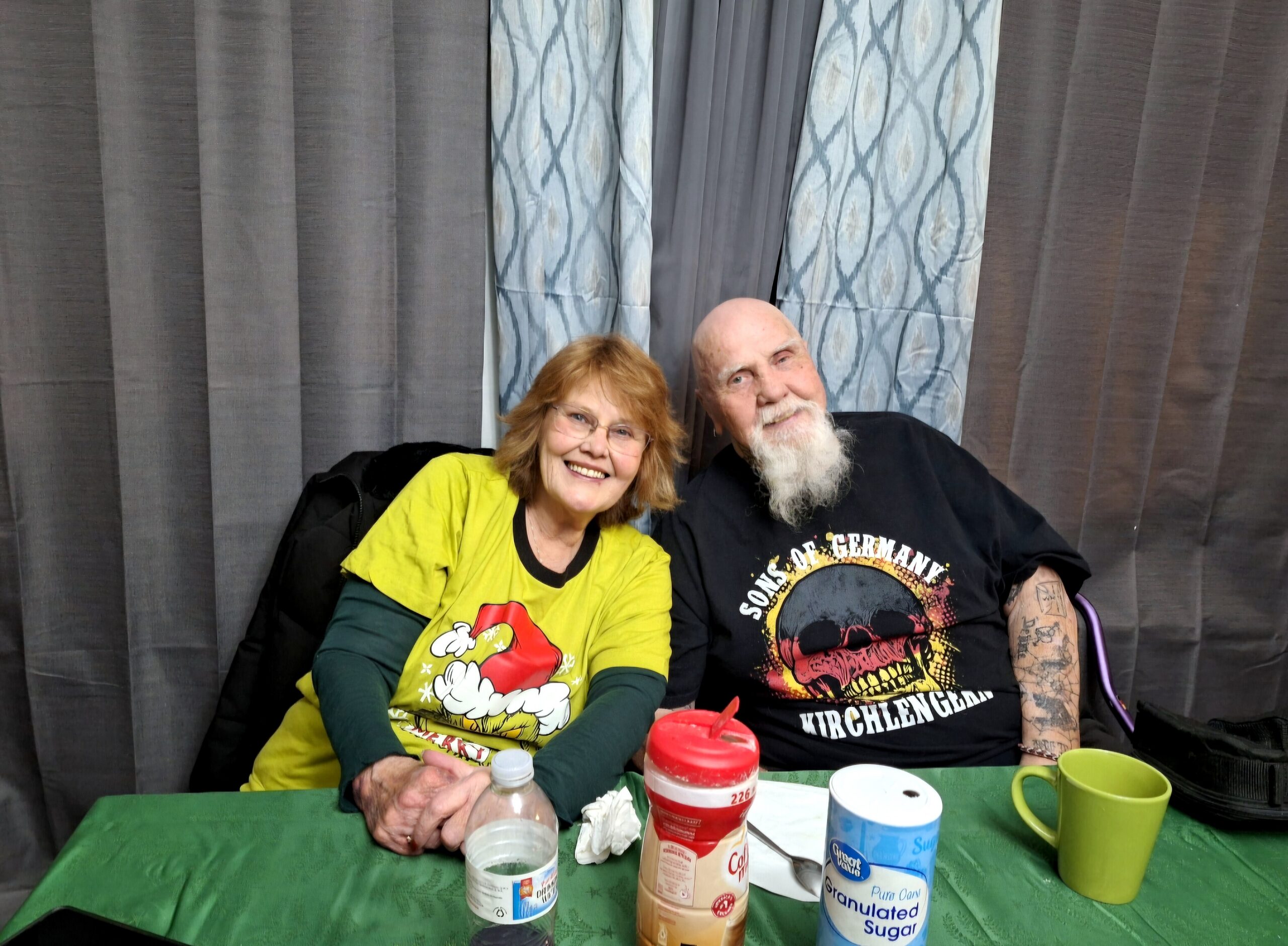 Smiling elderly couple seated at a green table with drinks, condiments, and cups, grey curtains in the background