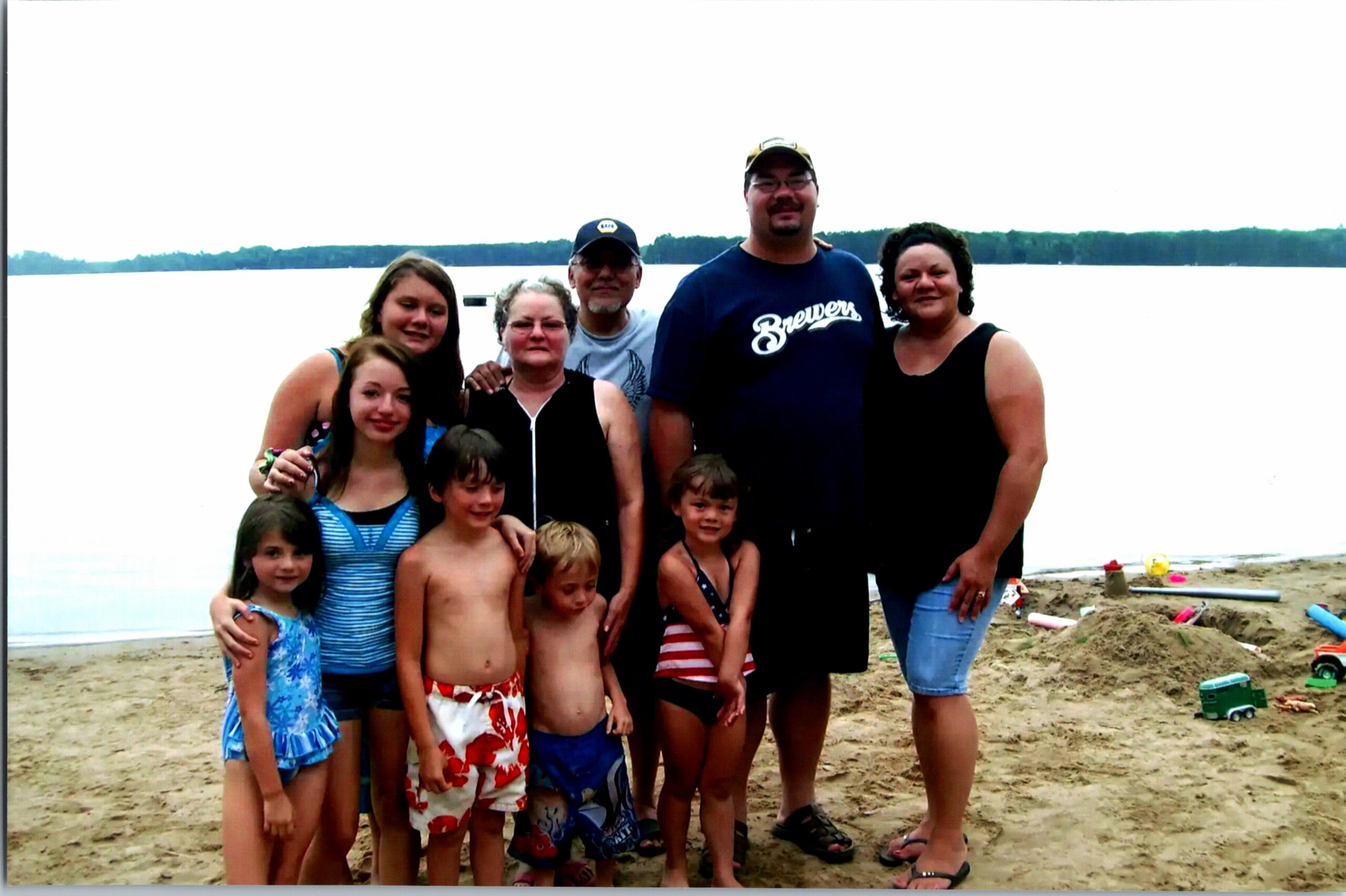 Group of adults and children posing together on a sandy beach with a calm lake in the background, smiling at the camera.