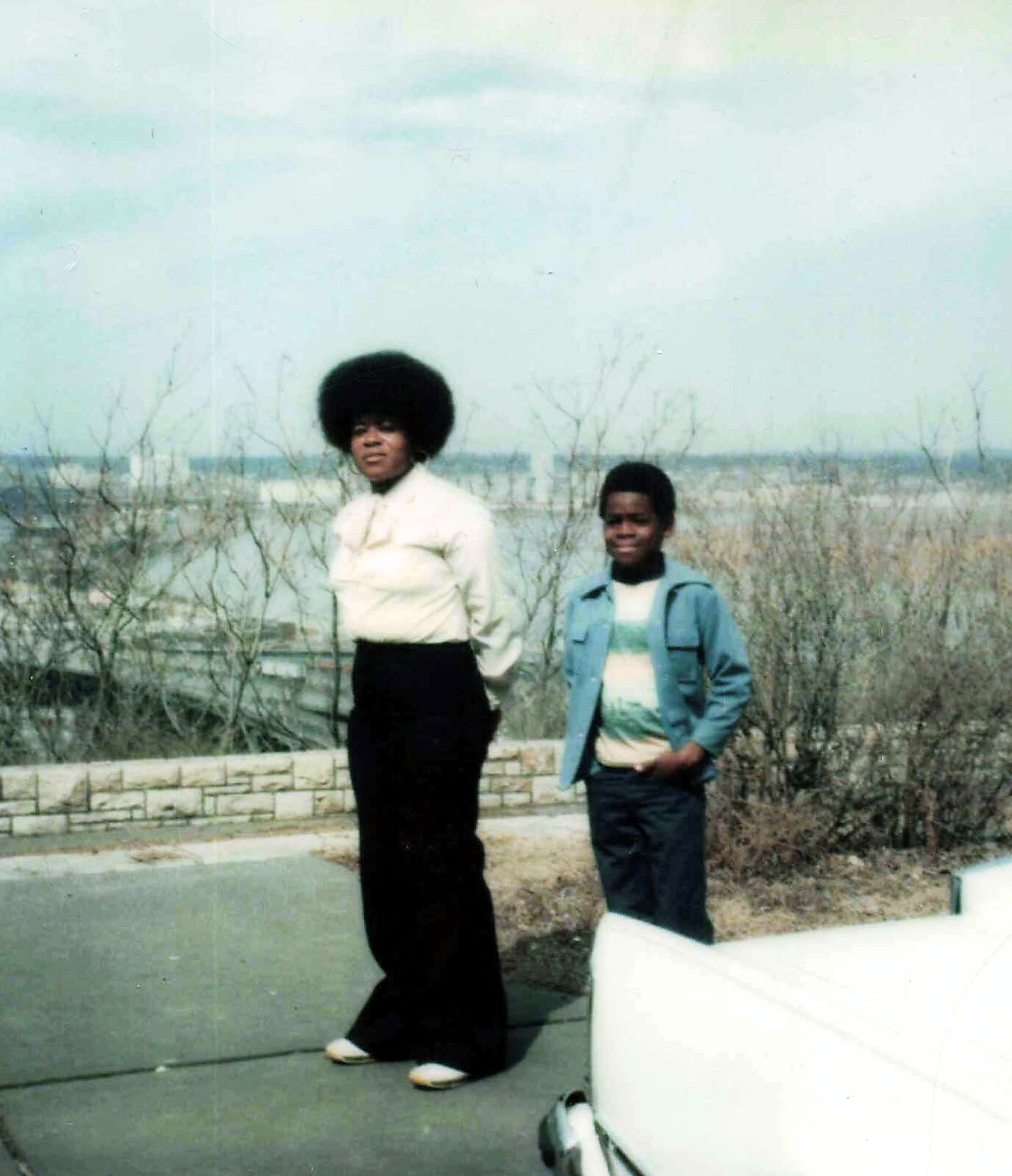 Two people posing outdoors near a low stone wall, a woman with a large afro and a boy in a denim jacket stand beside a white car.
