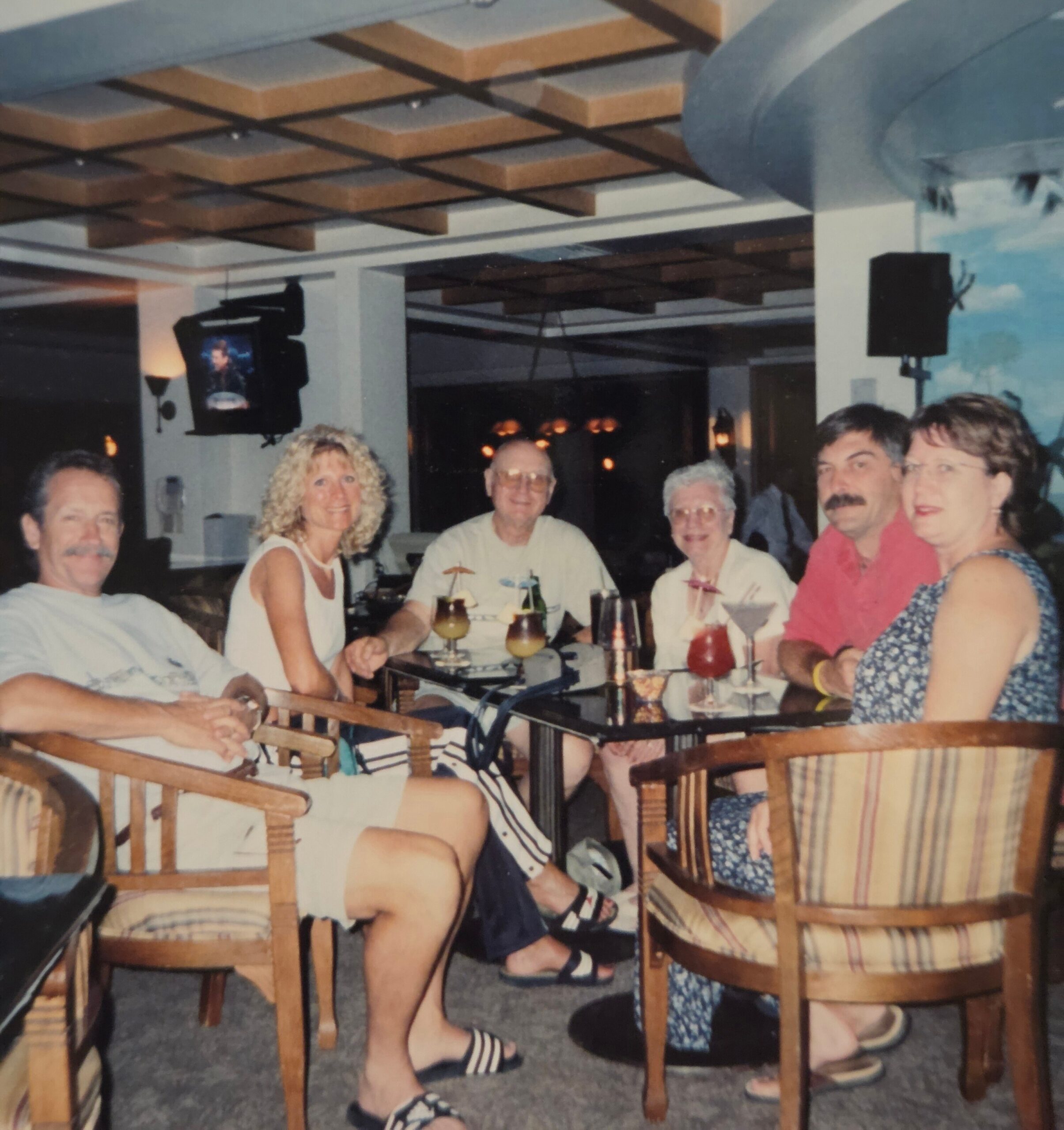 Group of adults sitting around a dining table with drinks, smiling at a casual restaurant setting.