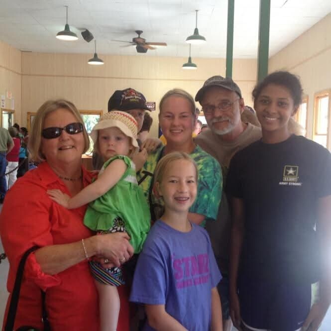 Smiling group of adults and children posing indoors for a casual photo; a woman in red holds a toddler in green, with two girls in front and others behind.