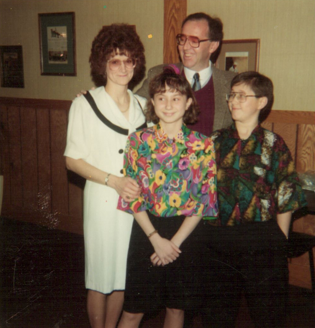Smiling family portrait indoors: a girl in a bright floral blouse stands in front with two women and a man behind them on a wood-paneled wall.
