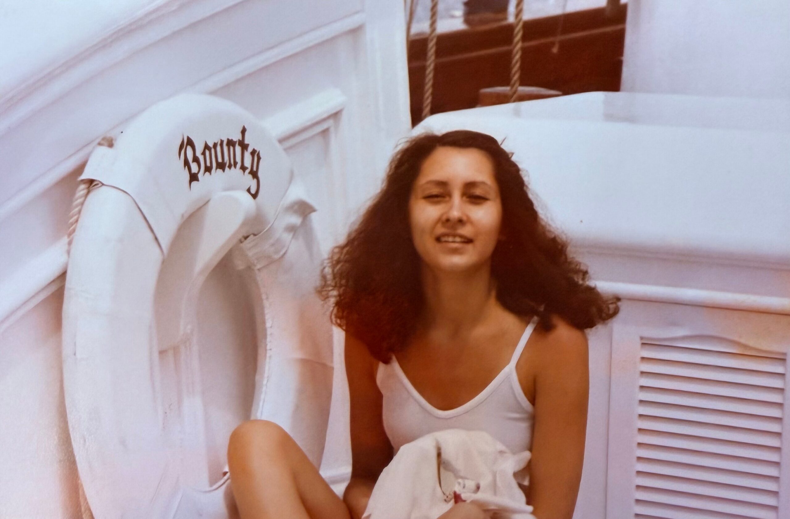 Young woman with curly hair in a white tank top sitting against a light wall, holding a folded blanket with a white lifebuoy behind her.