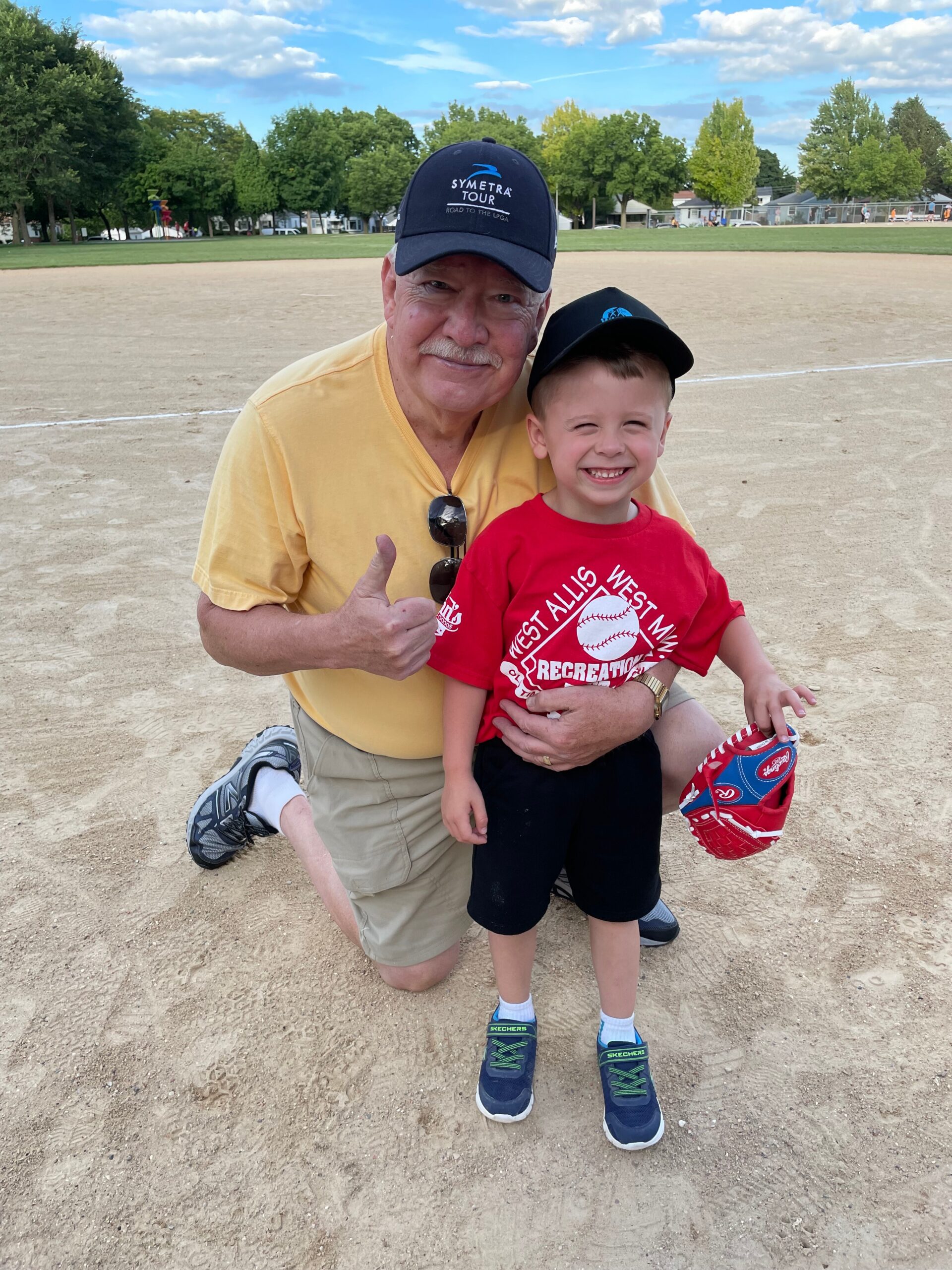Grandfather kneeling on a dirt baseball field, smiling and giving a thumbs-up with his young grandson wearing a red shirt and cap beside him.