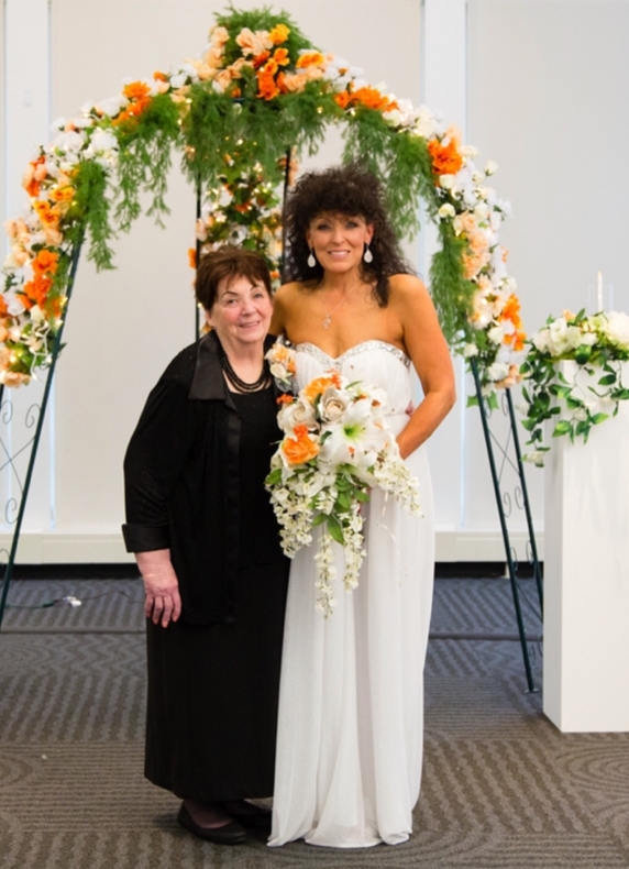 Bride in a white strapless gown holding a cascading orange-and-white bouquet, standing with an older guest under a floral arch at a wedding venue.