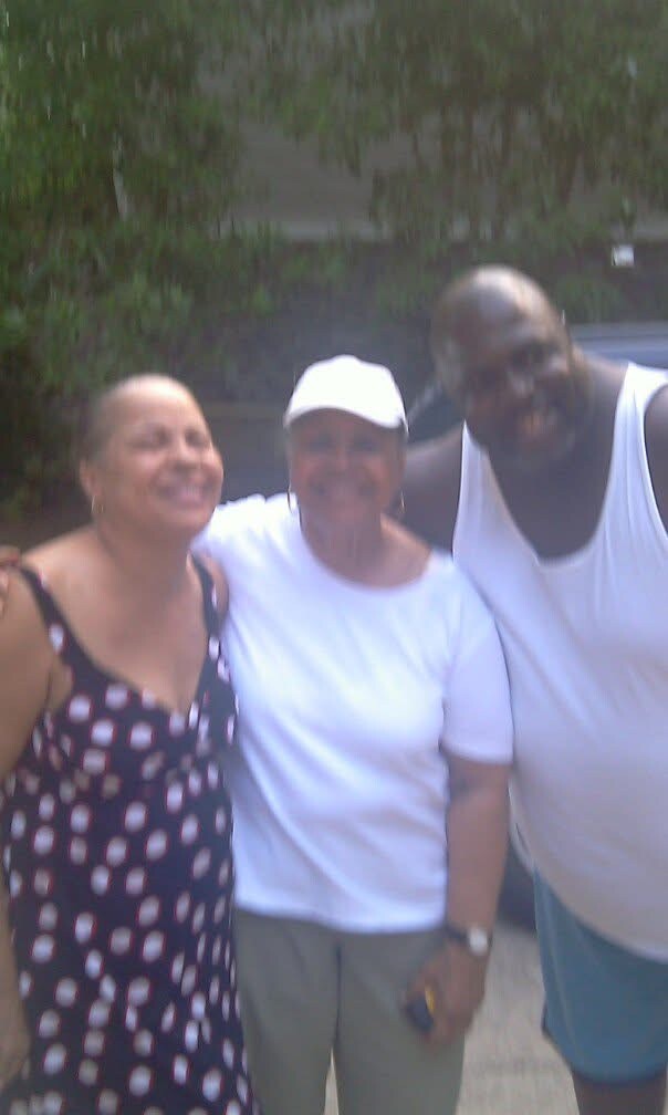 Three adults stand close together outdoors, smiling for the camera: a woman in a polka-dot dress on the left, a woman in a white cap in the middle, and a tall man in a white sleeveless shirt on the right.