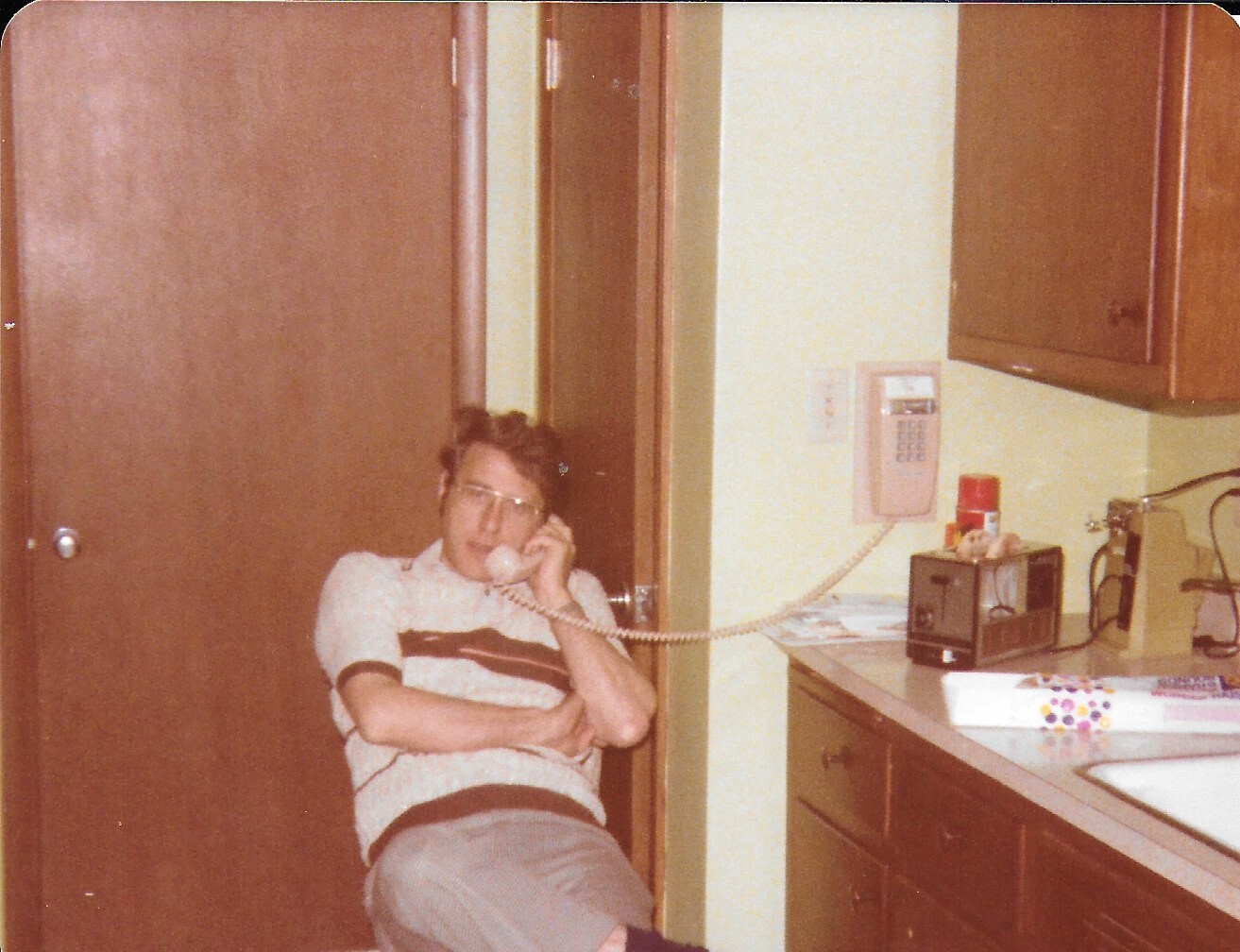 Man wearing glasses sits in a kitchen, talking on a corded wall phone mounted on the wall/near the counter.