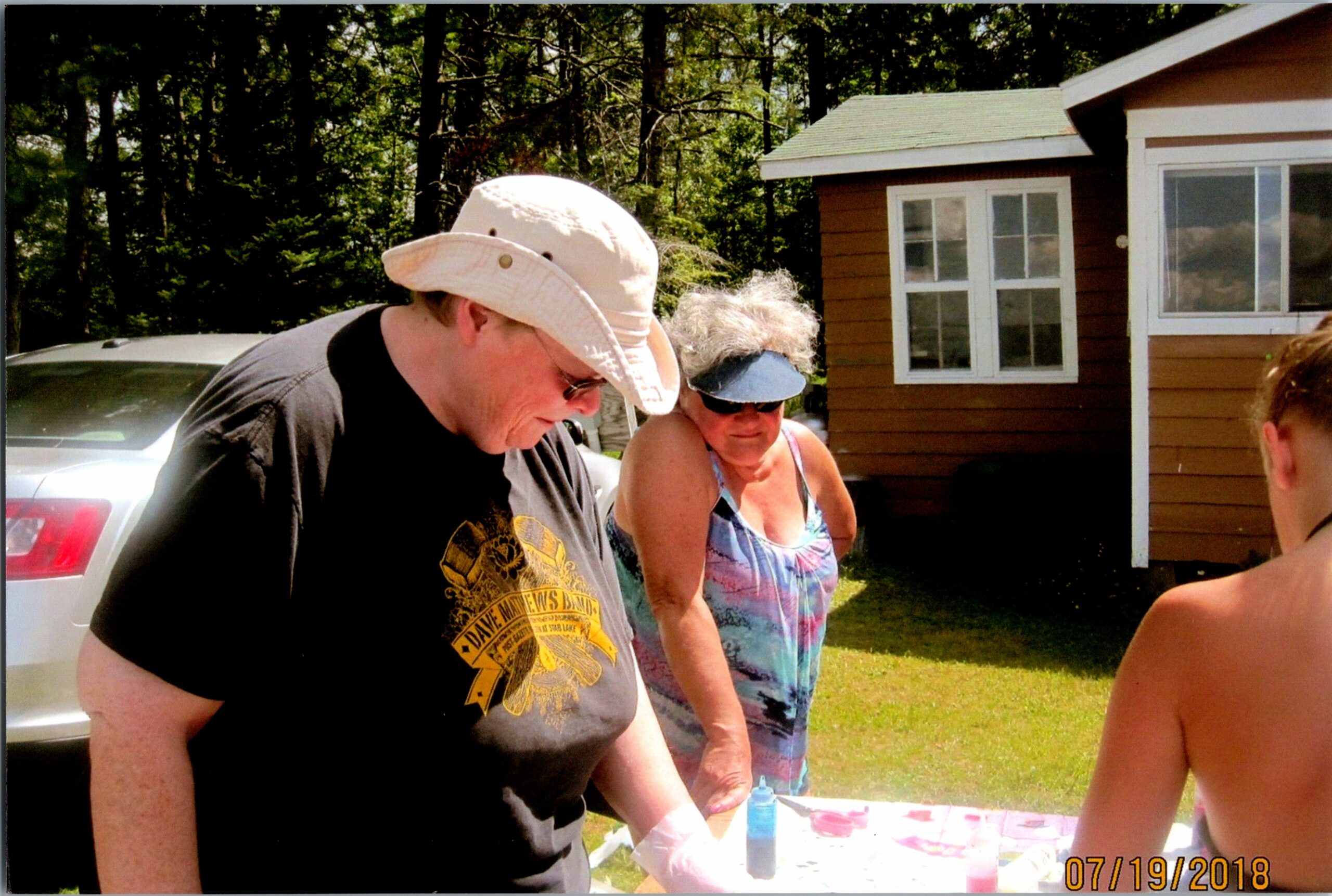 People gathered around a table outside, with paints and bottles visible, as a man in a light hat and a woman wearing a visor look down at the work. A house and trees are in the background. Date stamp 07/19/2018.