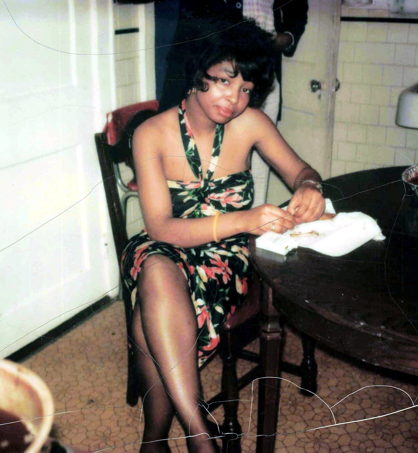 Woman in a tropical floral dress seated at a table, sewing fabric in a kitchen.