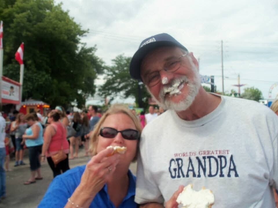 Smiling couple at an outdoor fair; man with frosting on his face wearing a 'World's Greatest Grandpa' shirt, holding cake pieces.