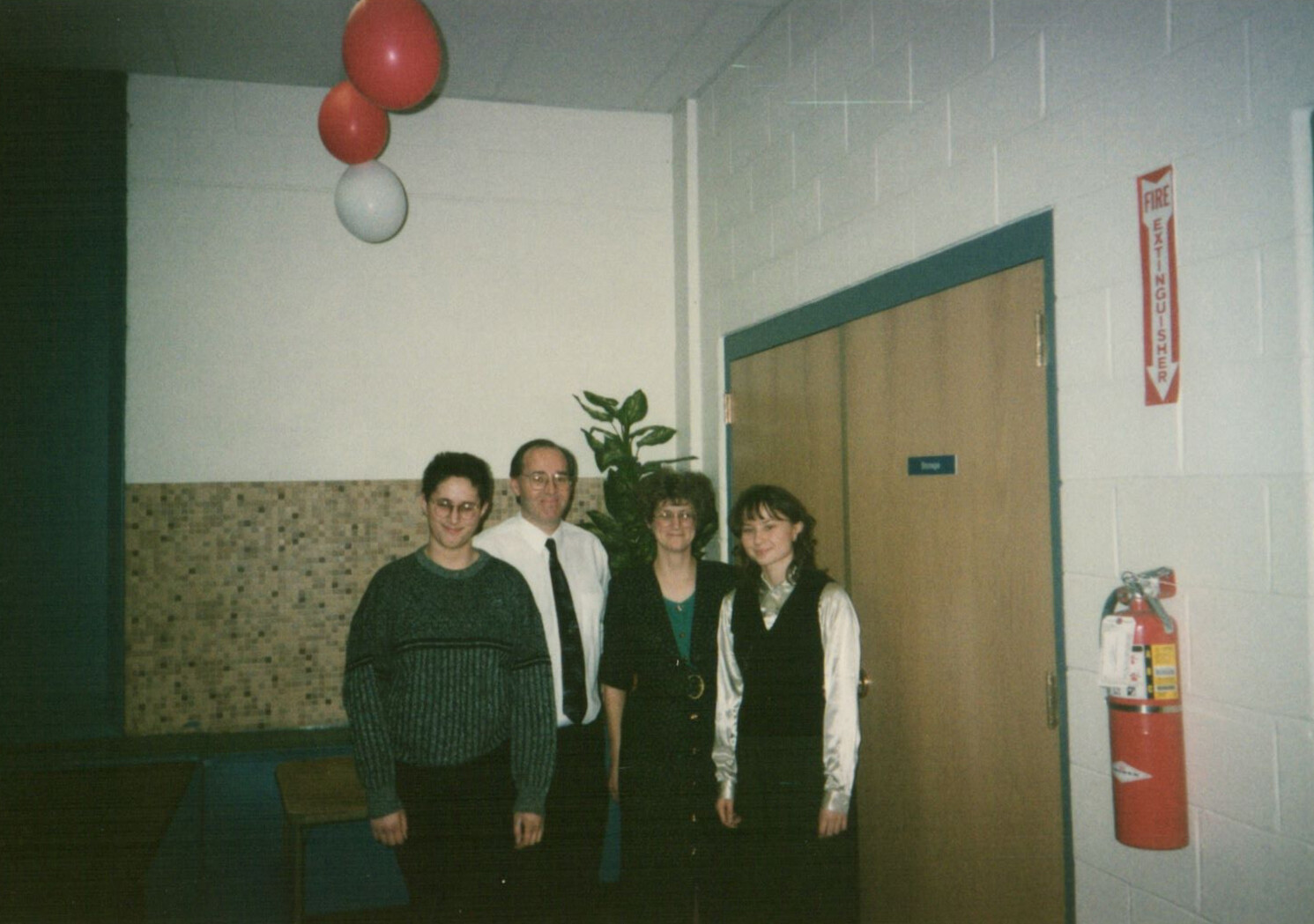 Four people pose in a hallway with balloons overhead, a potted plant behind them, and a fire extinguisher on the wall near a closed door.