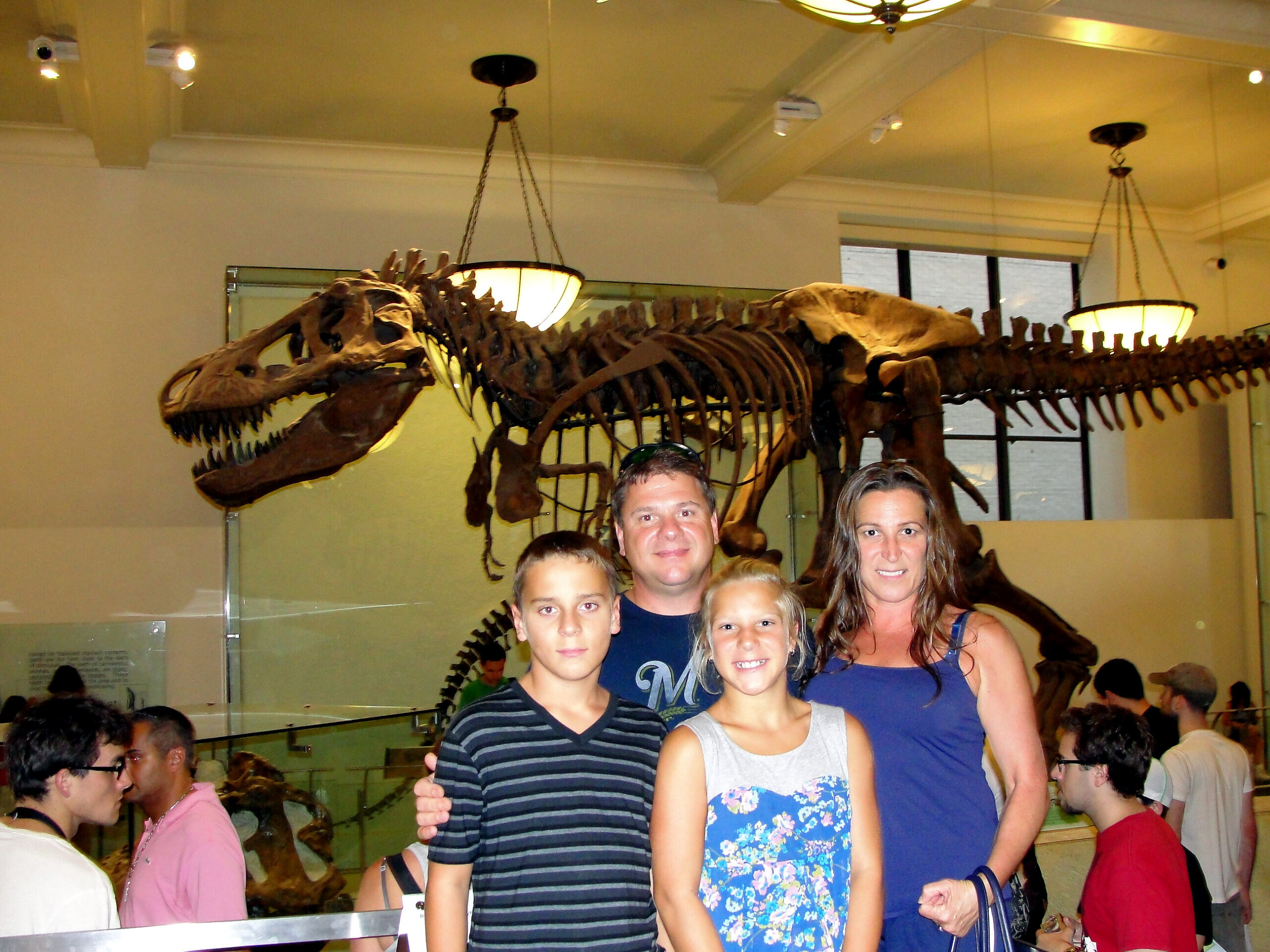 Group of people posing in front of a large hanging dinosaur skeleton in a museum gallery.