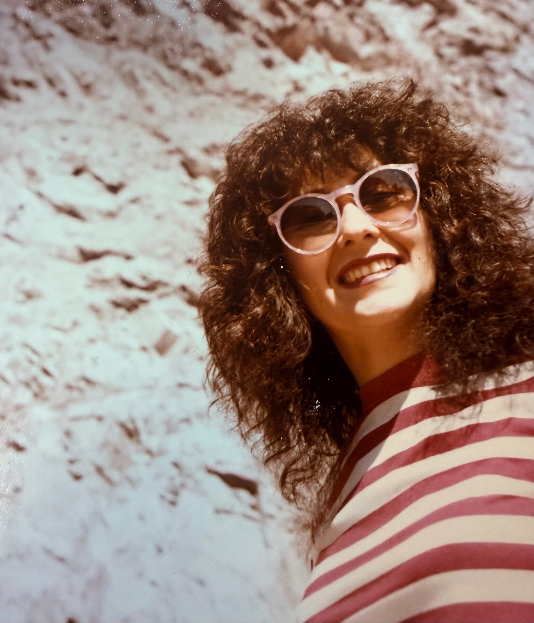 Smiling person with curly hair wearing large white sunglasses and a red-and-white striped shirt, outdoors against a rocky background.