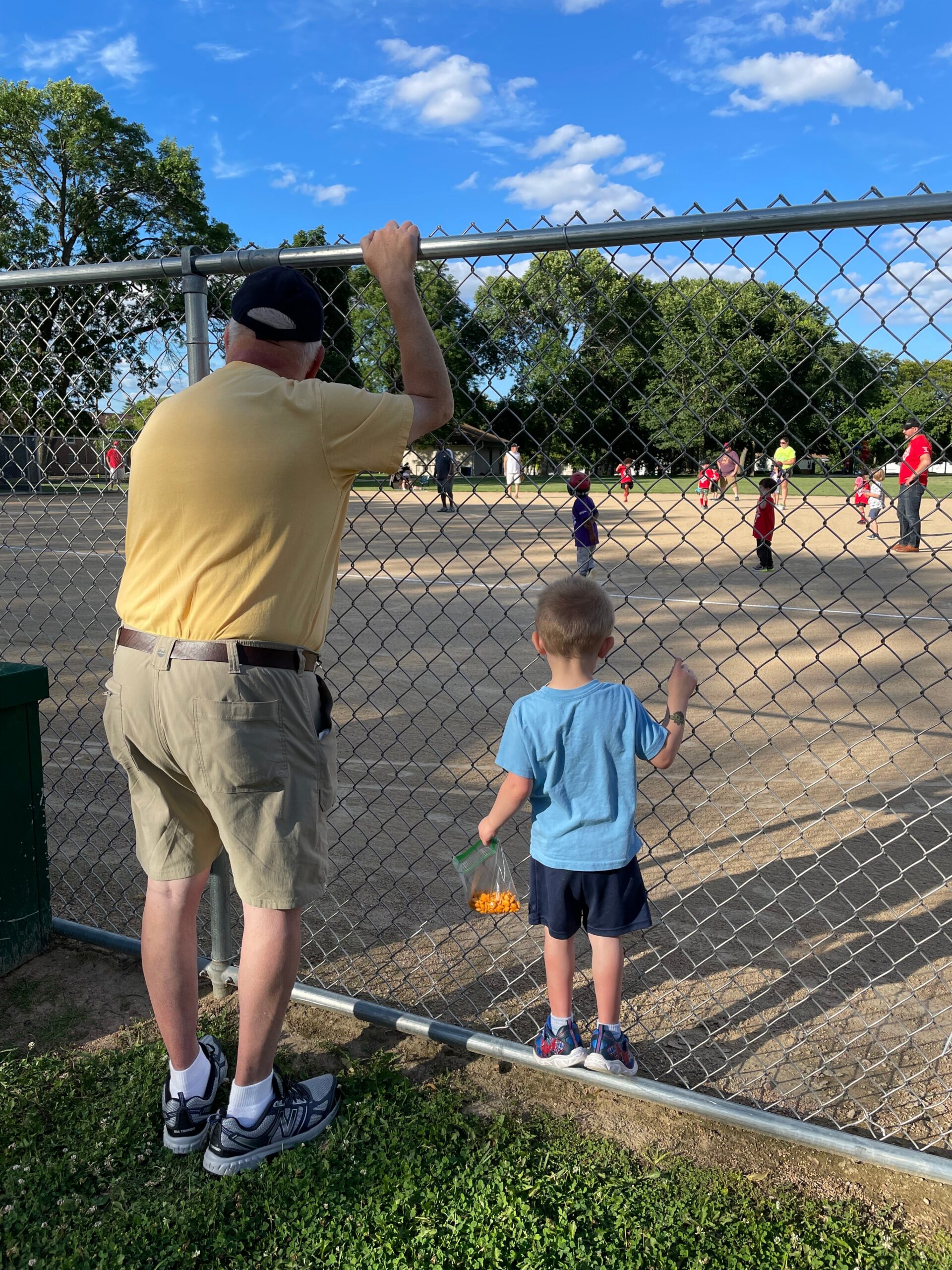 Older man in khaki shorts and a yellow shirt stands at a chain-link fence watching kids play baseball on a sunny day.