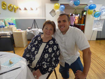 Smiling elderly woman seated beside a younger man at a birthday party; balloons and a cake table visible in the background.