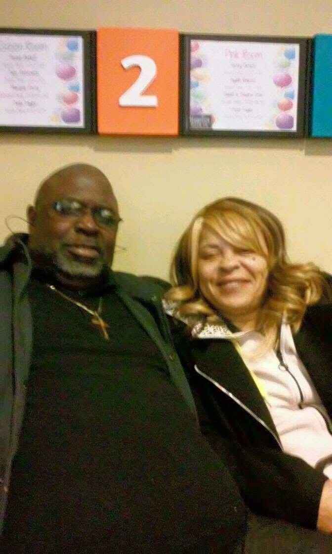 Smiling couple seated side by side in a waiting area, with a colorful wall display featuring a large orange '2' behind them.