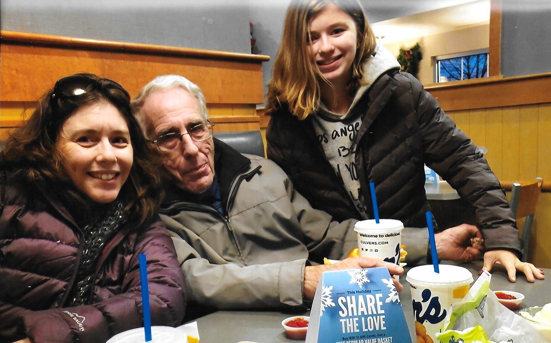 Three people sit at a cafe table, smiling at the camera, with drinks and a blue promotional card on the table.