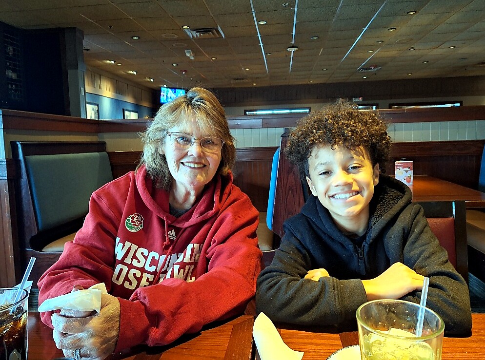 Two people seated at a restaurant booth smiling at the camera: an older woman in a red Wisconsin hoodie and a smiling young boy in a dark hoodie with drinks on the table