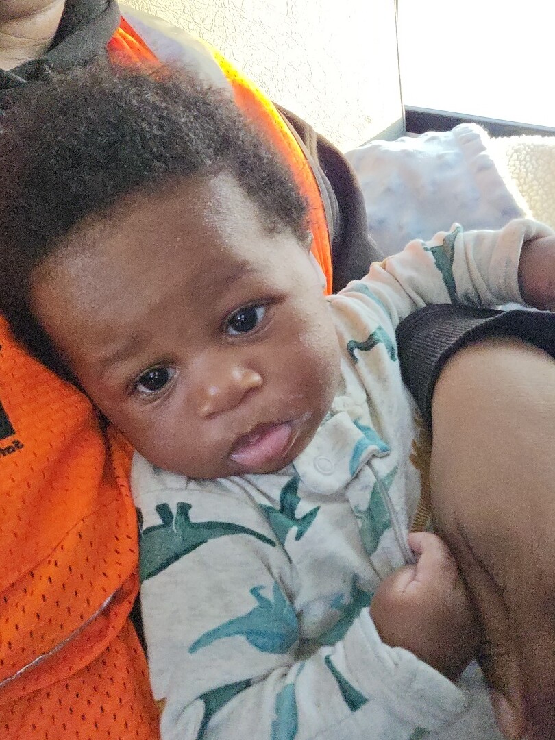 Close-up of a baby resting in an adult's arms, wearing a white onesie with teal dinosaurs, looking toward the camera in a bright setting.