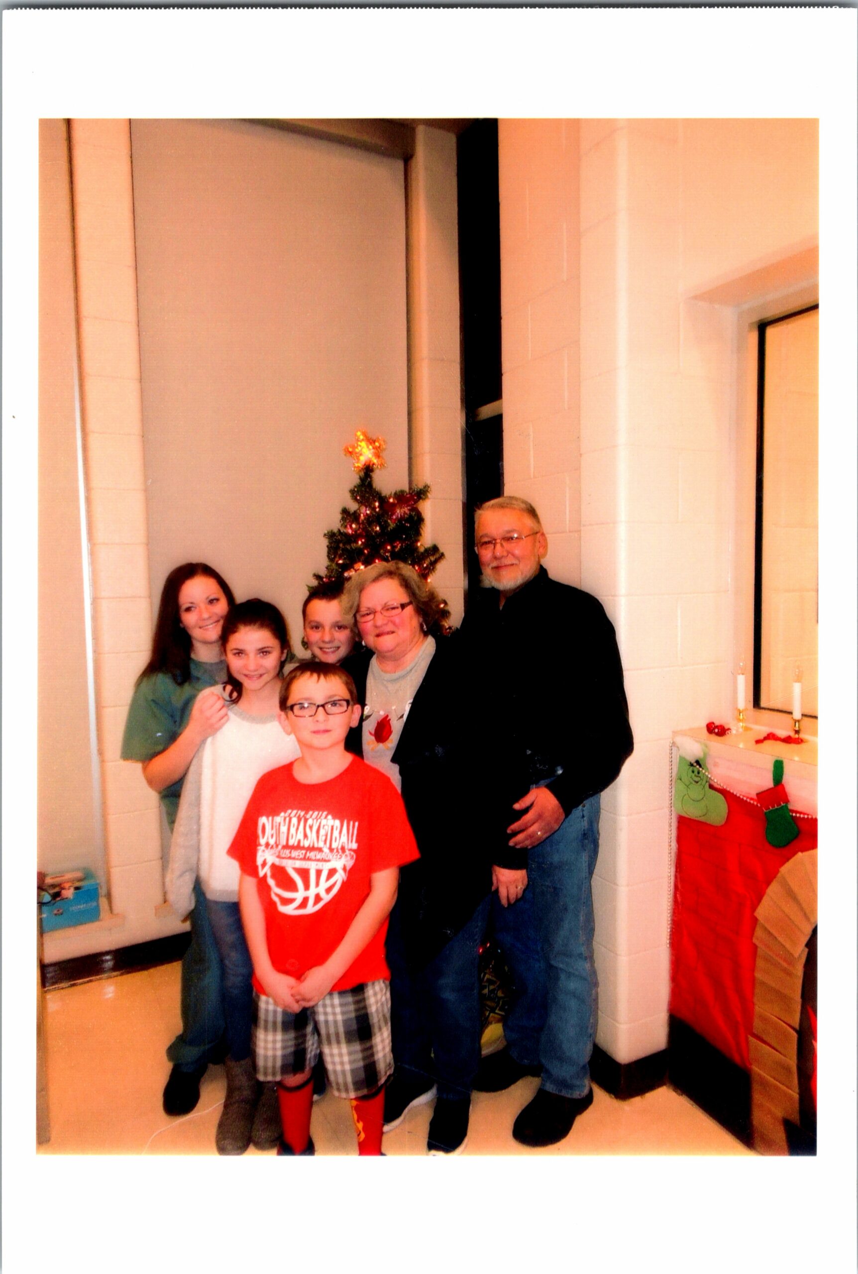 Group of seven people posing for a Christmas photo indoors, with a small decorated tree and stockings visible in the background.