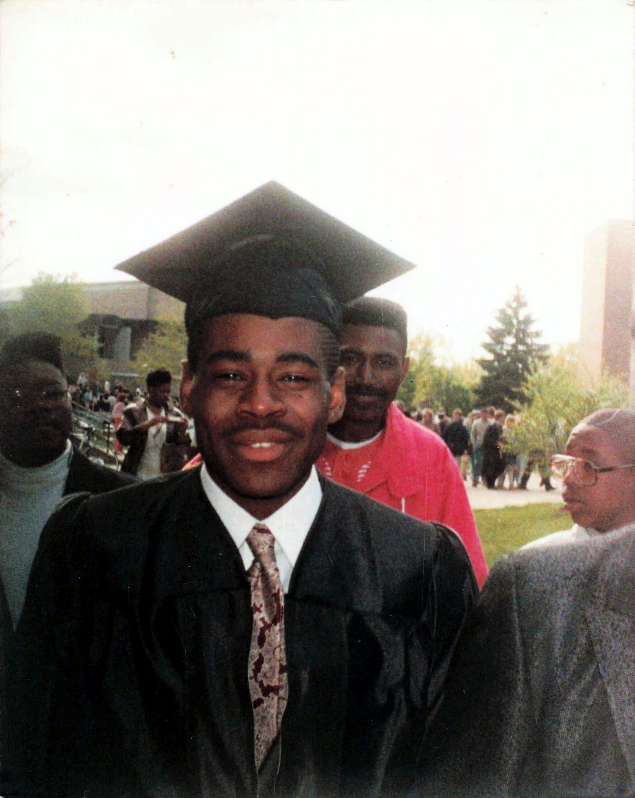 Young man in a black graduation gown and cap smiles at the camera during an outdoor graduation ceremony.