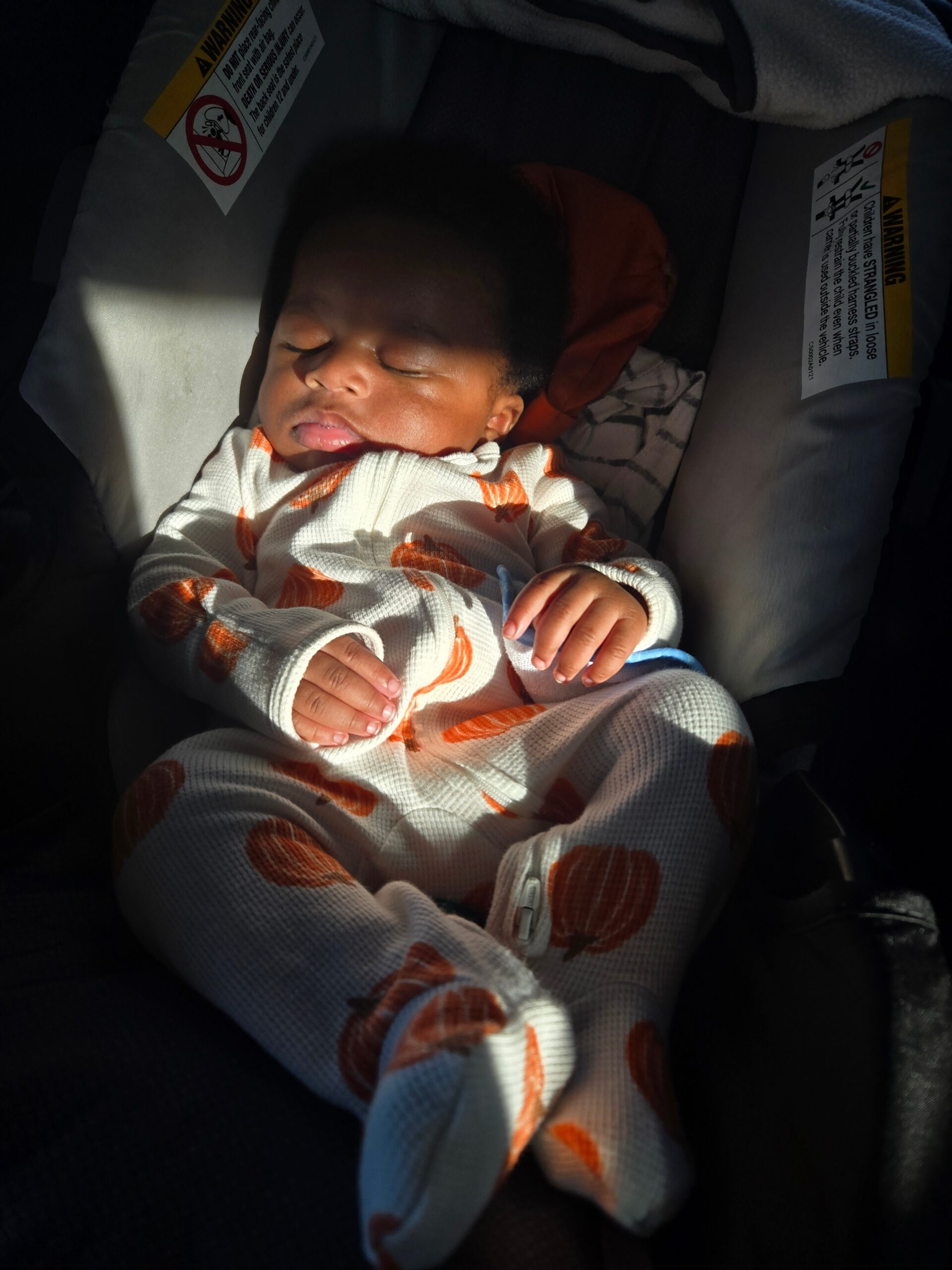 Sleeping baby in a car seat wearing a white onesie with orange shapes, eyes closed.
